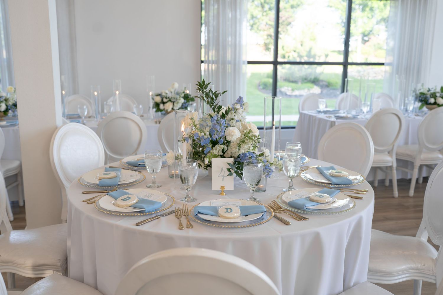 Elegant wedding reception table with white linens, blue accents, floral centerpiece, and gold flatware in a bright venue.