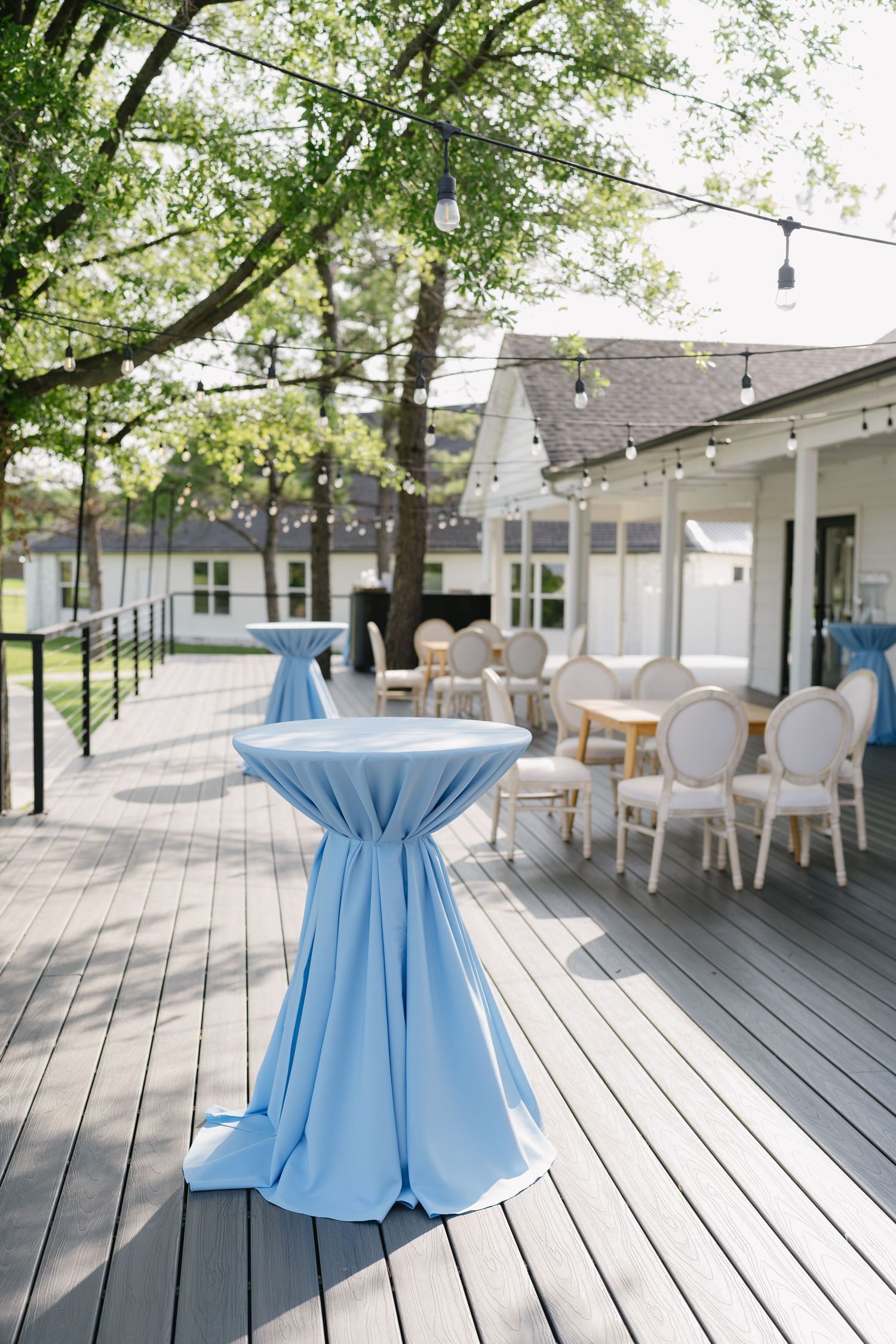 Elegant outdoor deck with light blue cocktail tables and white chairs, string lights, and lush green trees at an event venue.