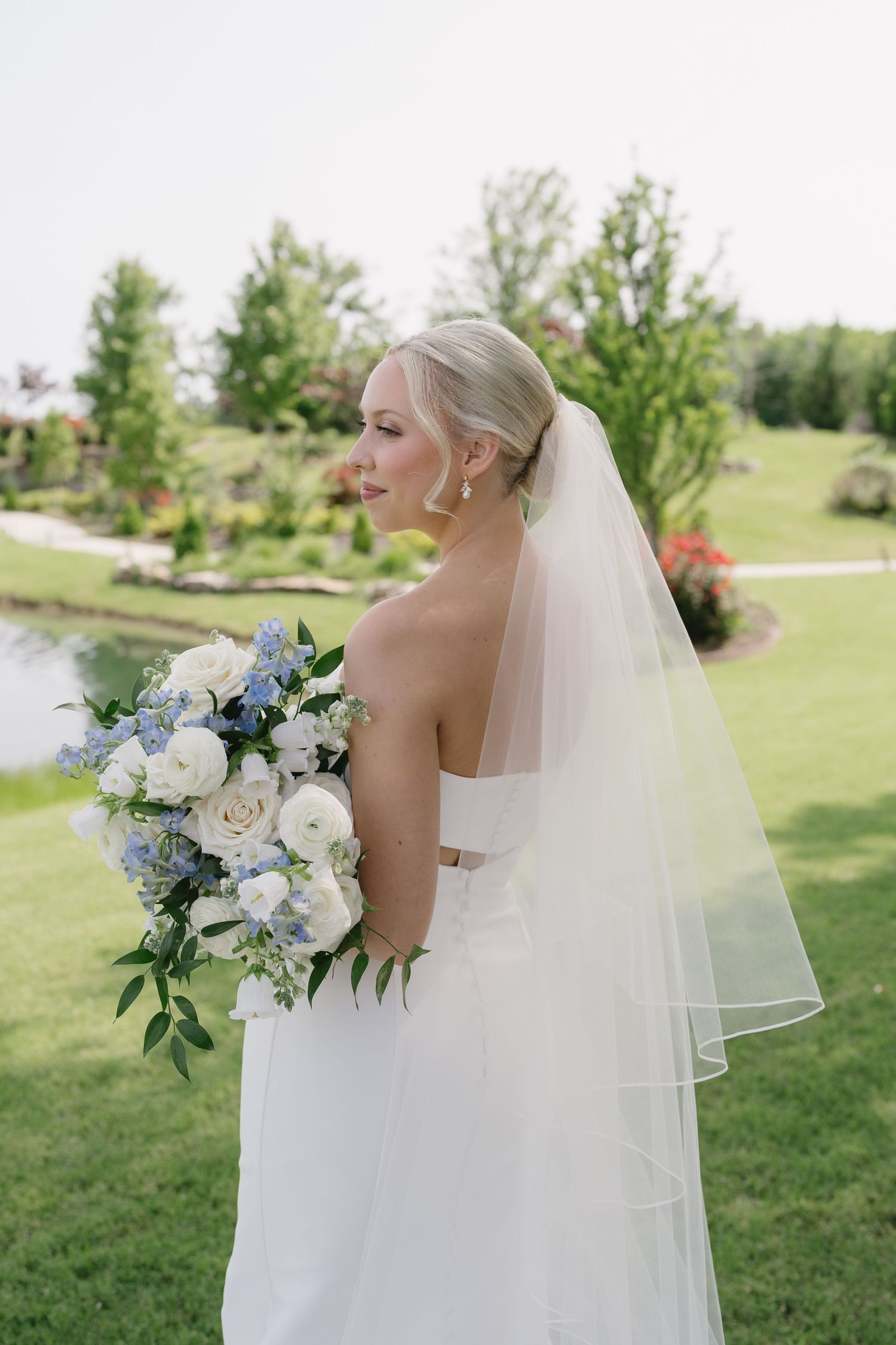 Bride in strapless white gown and veil holds blue and white bouquet, posing in lush garden near pond.