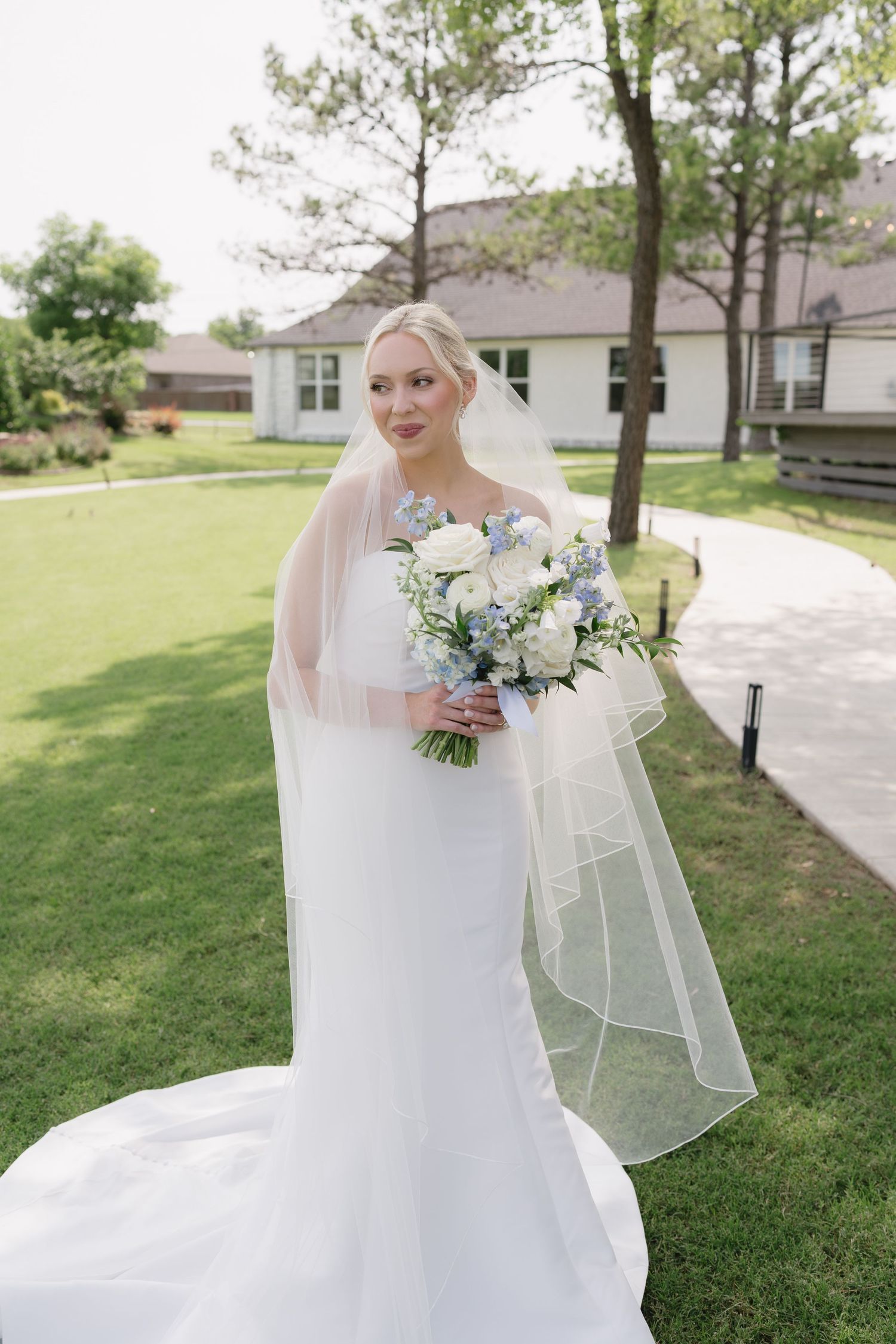 Elegant bride in white gown and veil holding blue and white bouquet standing on green lawn outside.