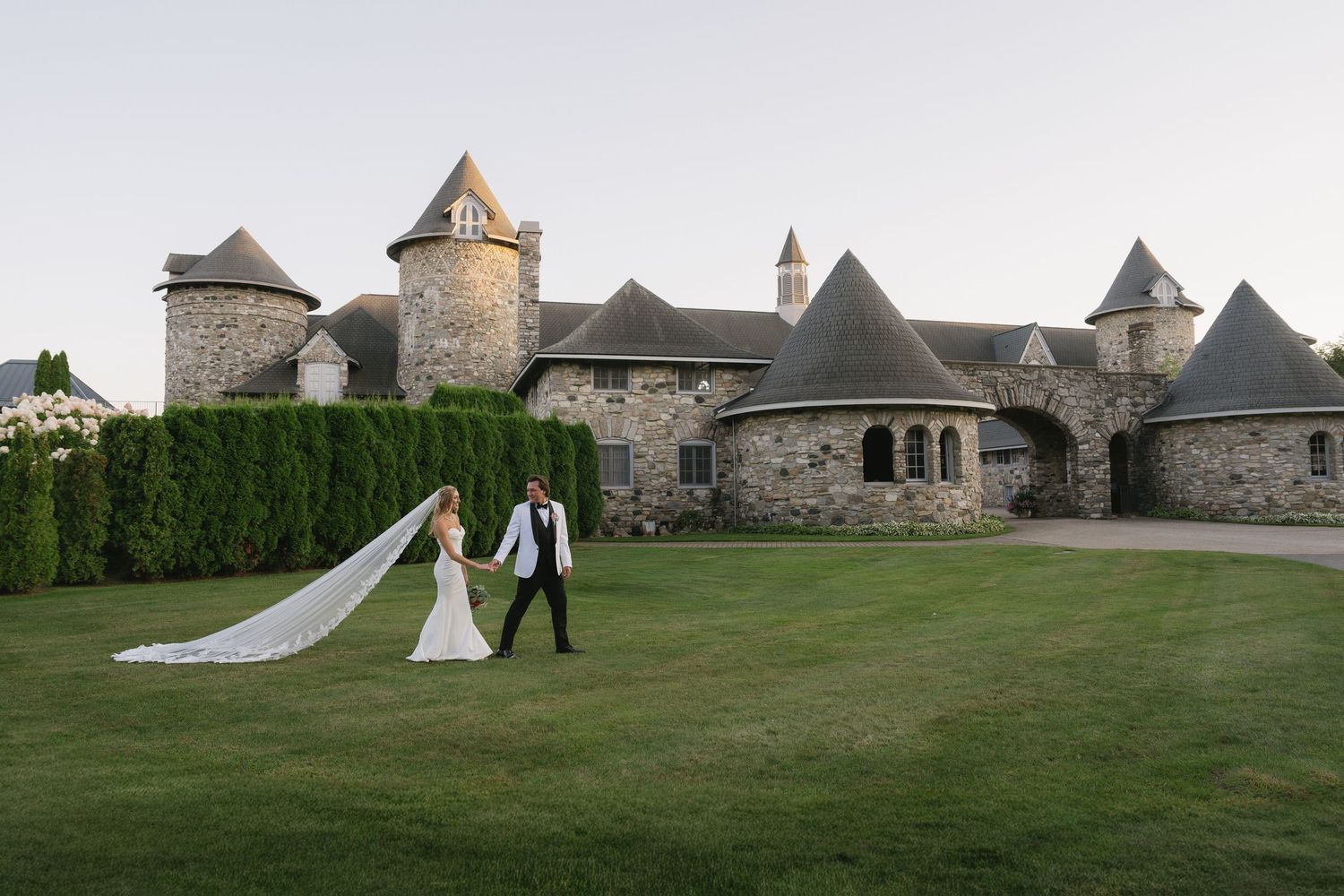 Bride and groom walk hand-in-hand across lush green lawn before a stunning stone castle wedding venue.