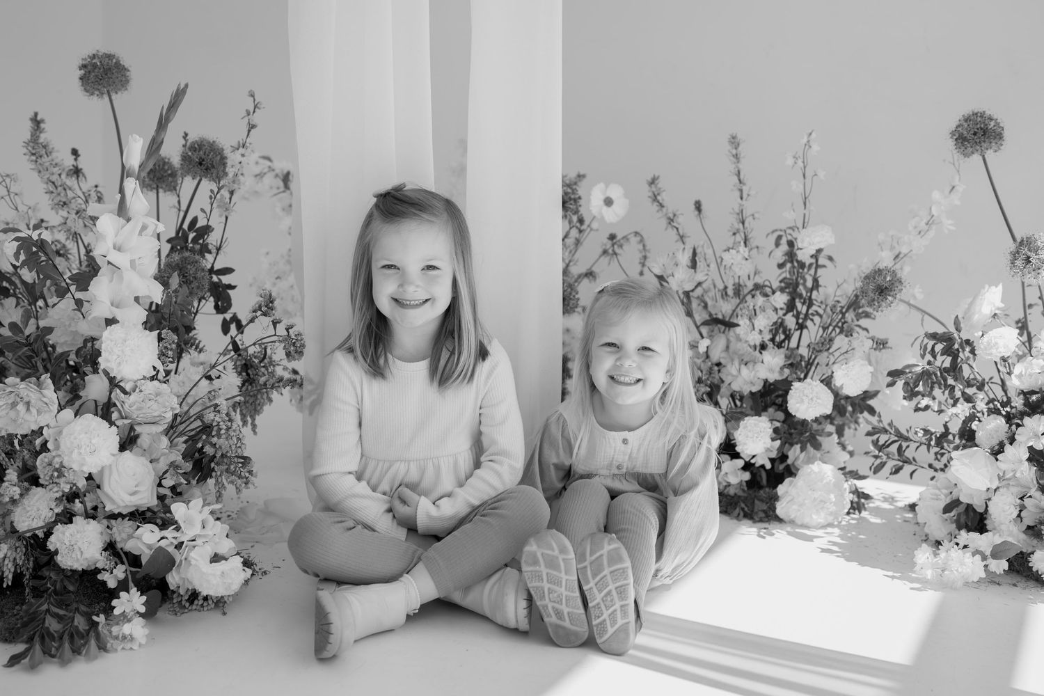 Black and white studio portrait with siblings sitting together among floral arrangements.