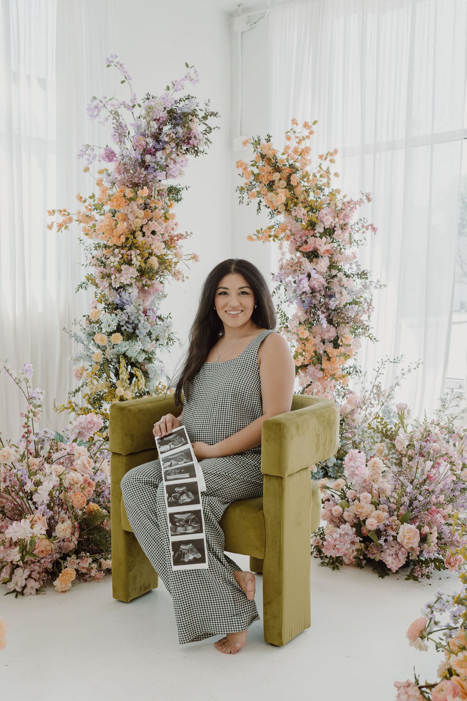 Expectant mother sits in olive chair holding ultrasound photos surrounded by floral arrangements.