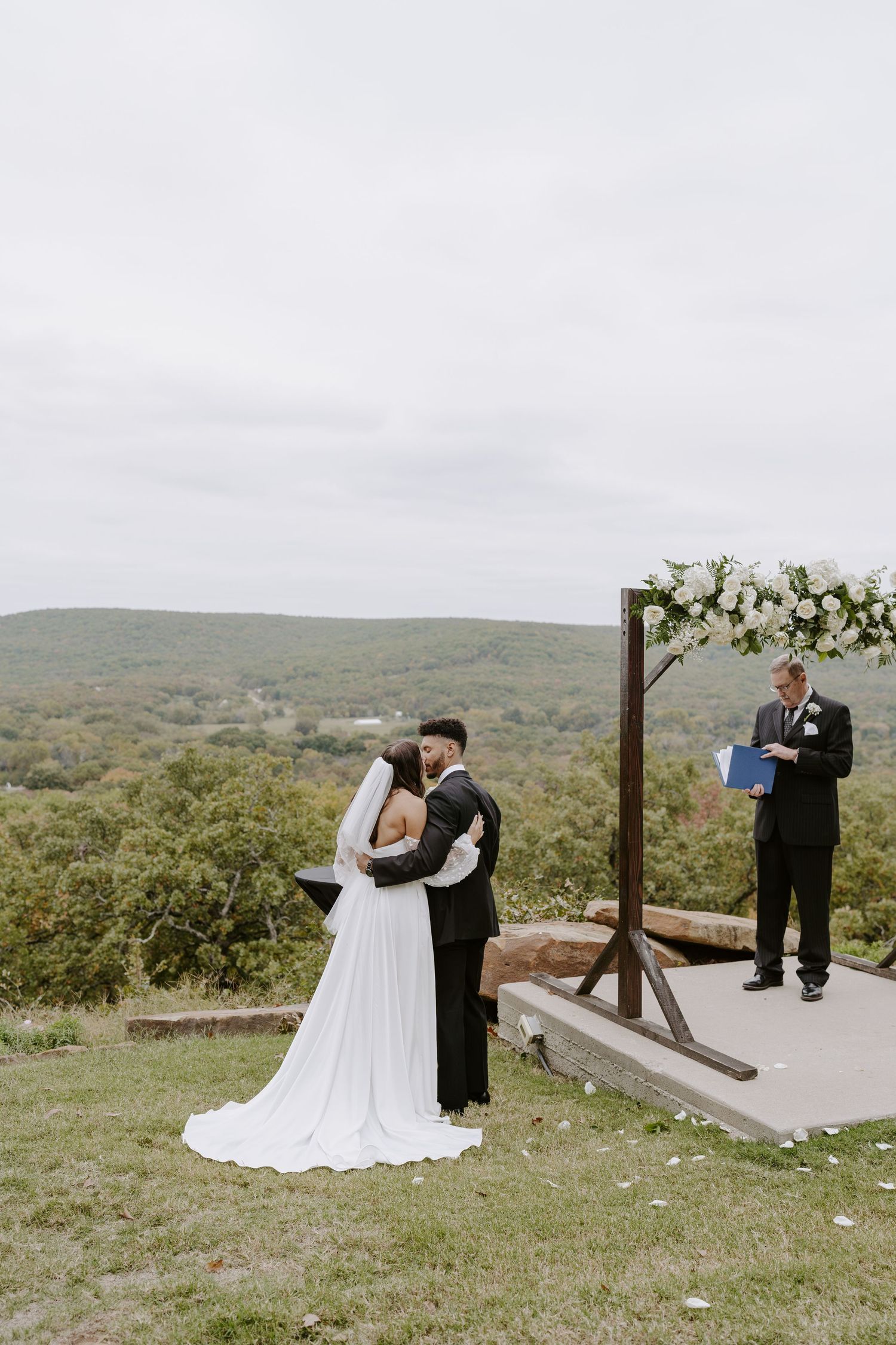 Wedding couple exchanging vows under wooden arch decorated with white flowers on scenic overlook.