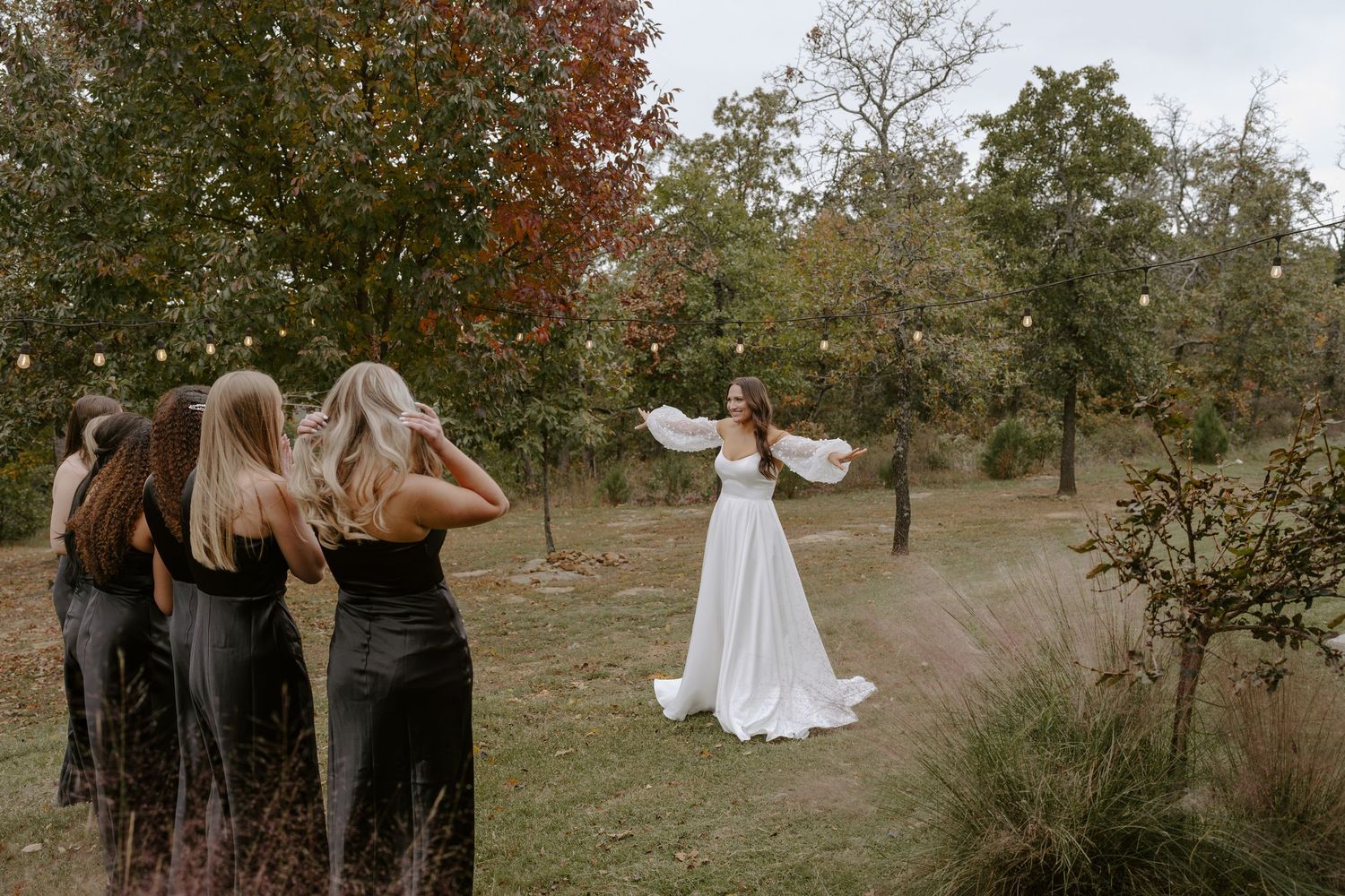 Wedding party in dark dresses observes as person in white gown twirls on grassy hillside.