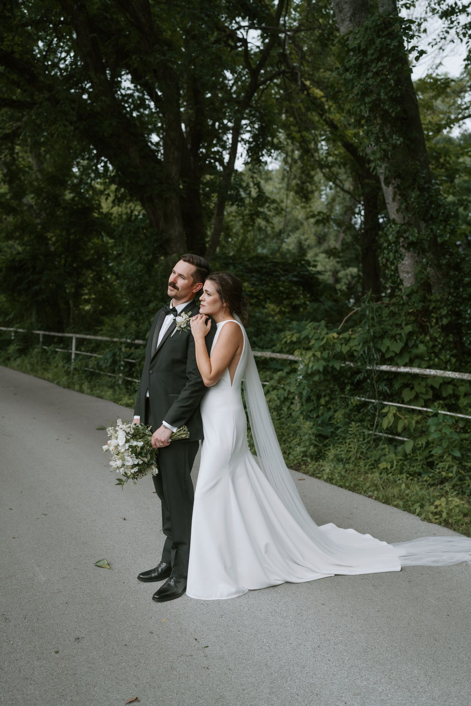 Wedding couple stands together on a winding path with overhanging trees and natural greenery.