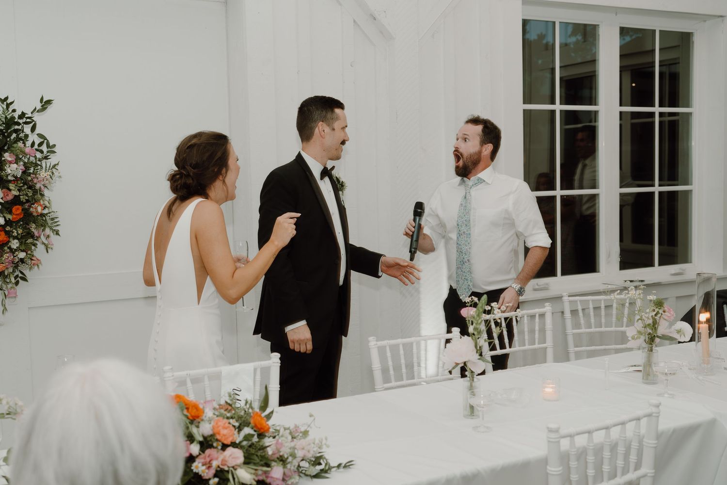 Wedding guests mingle and socialize in a bright white reception space with floral arrangements.