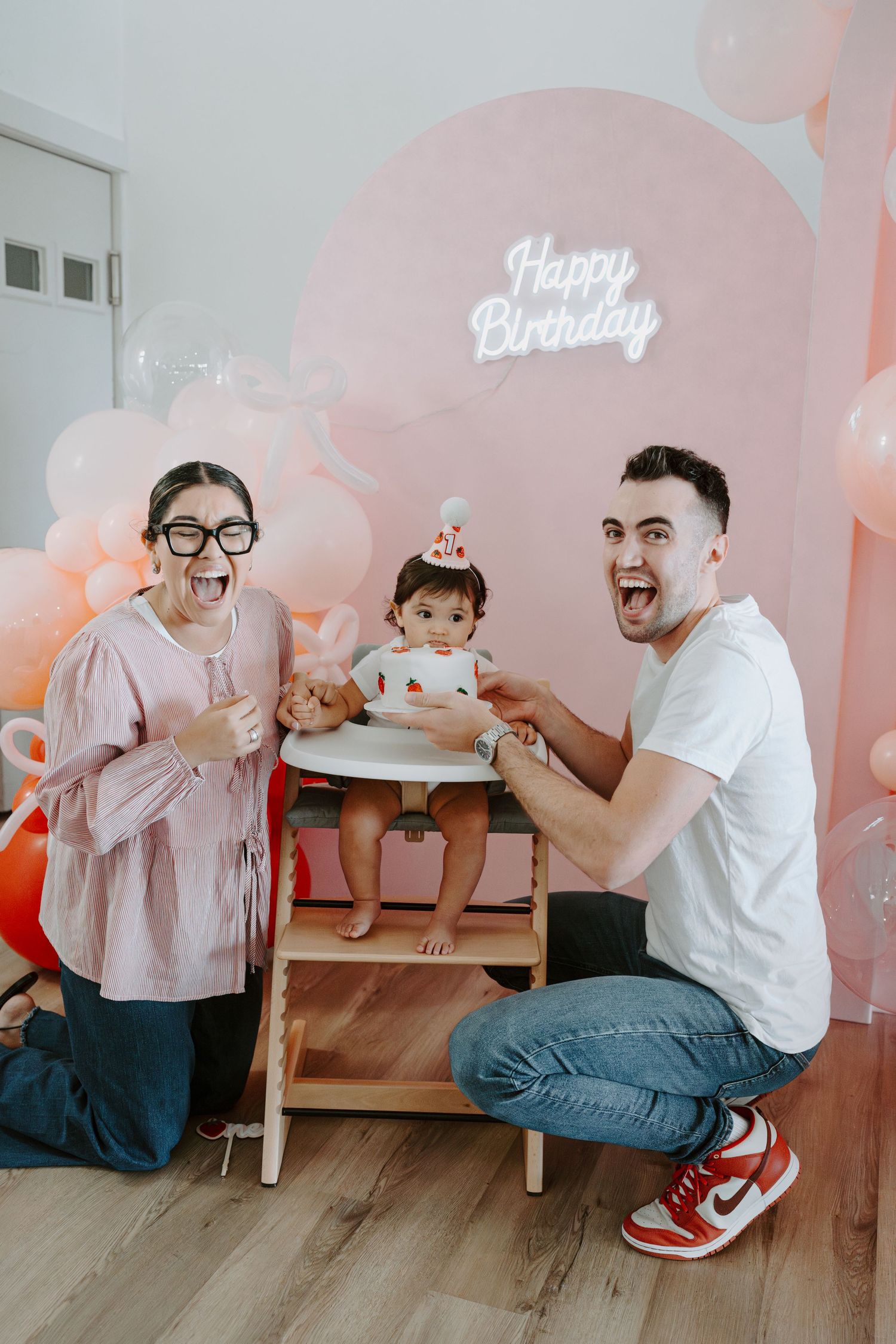 Intimate birthday celebration with family members gathered around small table with decorative pink background.
