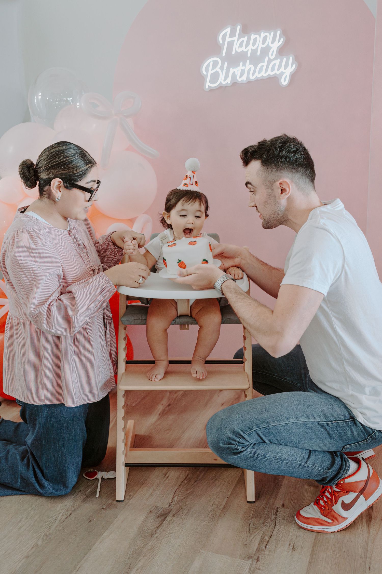 Family celebrating baby's birthday with cake in front of pink neon Happy Birthday sign.