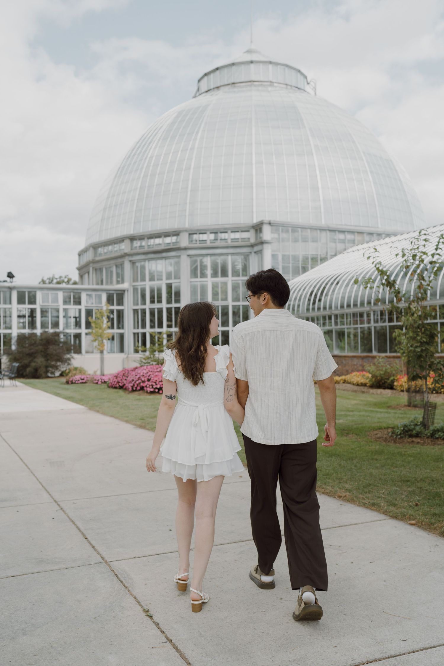 A couple walks together on a concrete path leading to a large glass greenhouse dome.