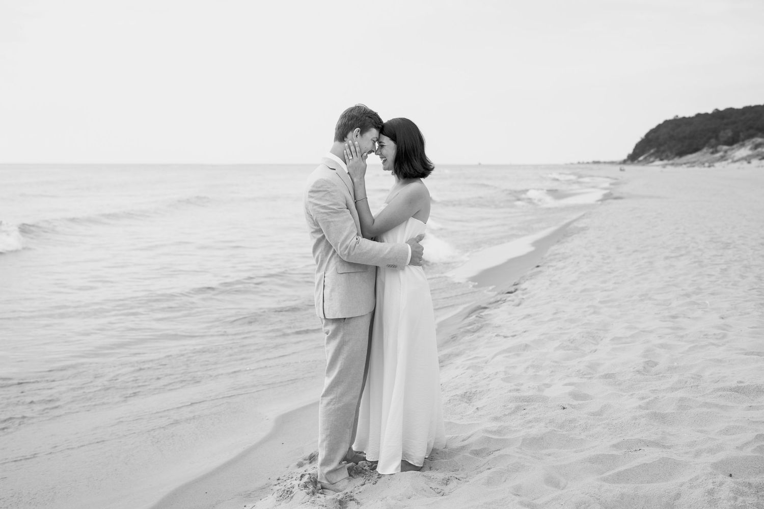 A romantic beach wedding couple embraces in black and white photography on a sandy shoreline.