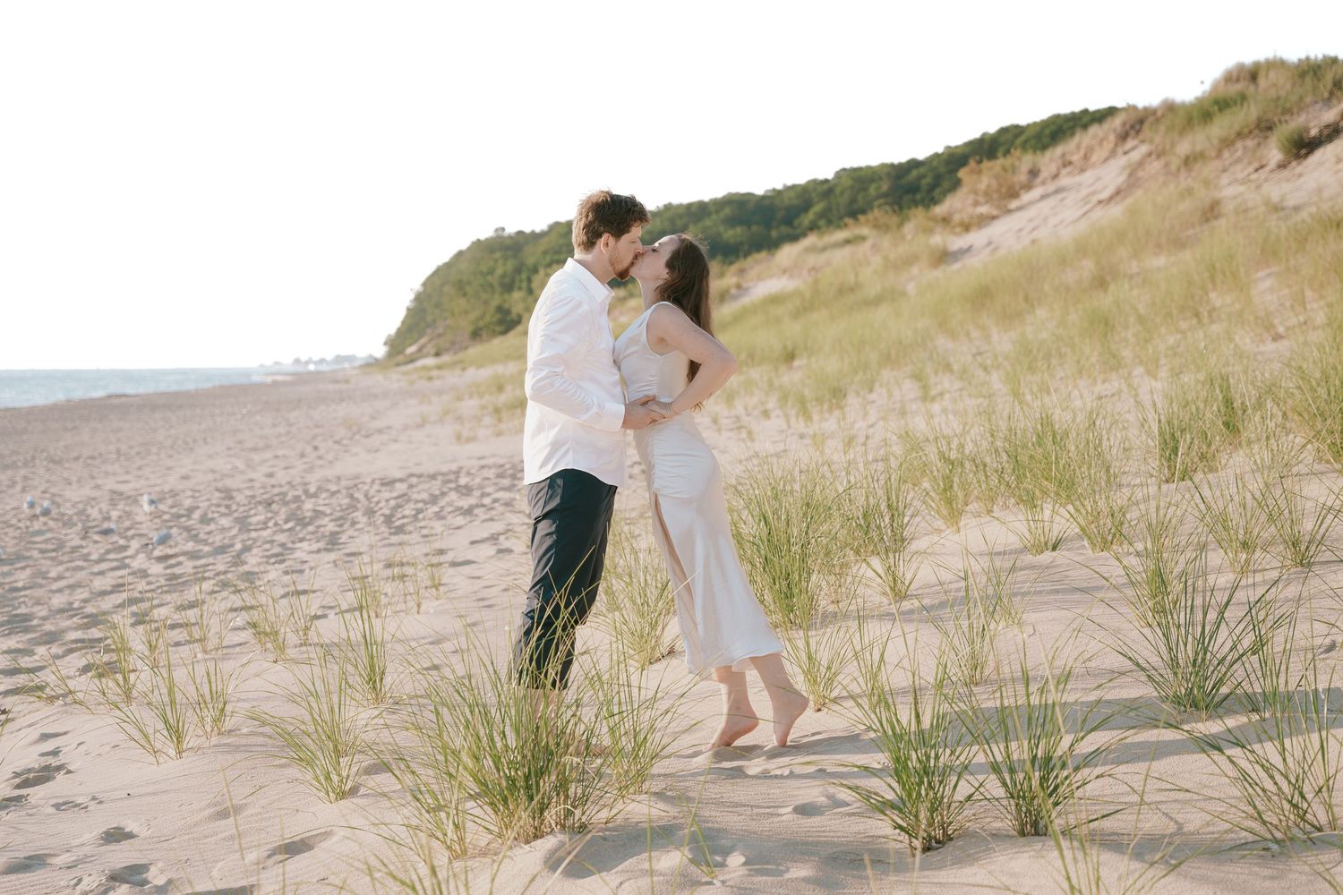A couple shares an intimate moment on a beach with coastal grasses and dunes in the background.