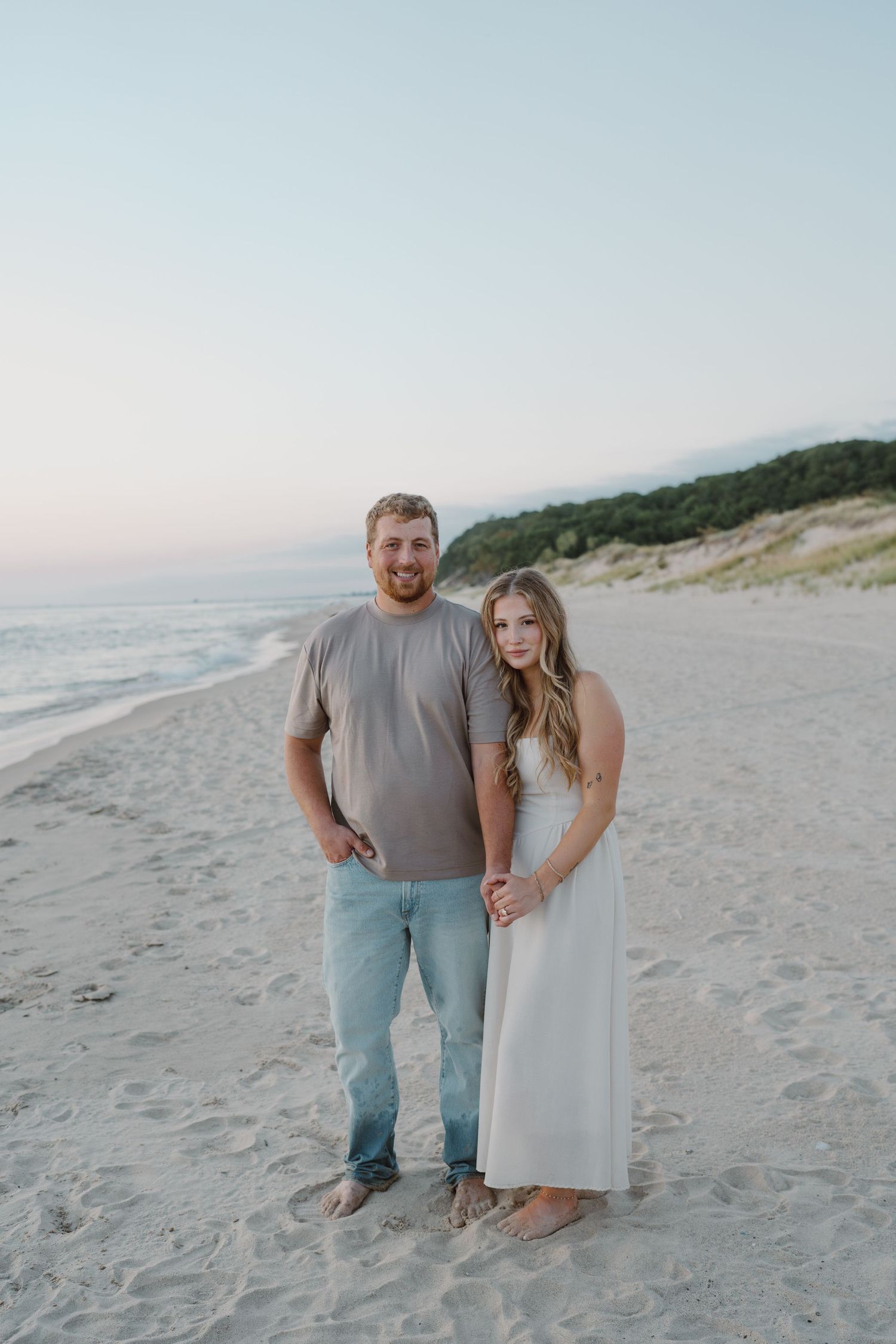 Couple poses together on sandy beach with ocean and dunes in background.