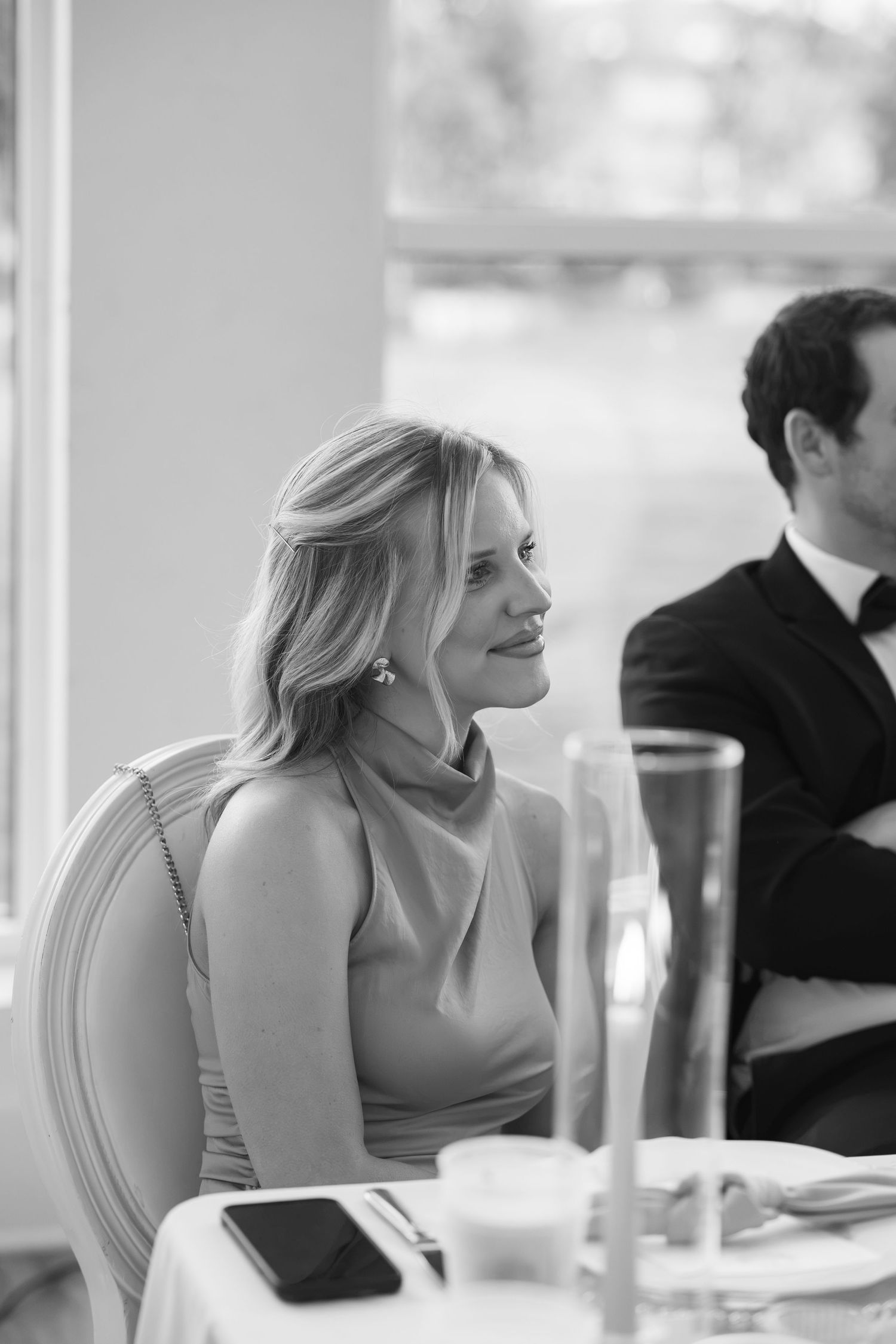 Elegant blonde woman smiling at formal dinner event, black and white photo, wearing sleeveless dress with pearl earrings.