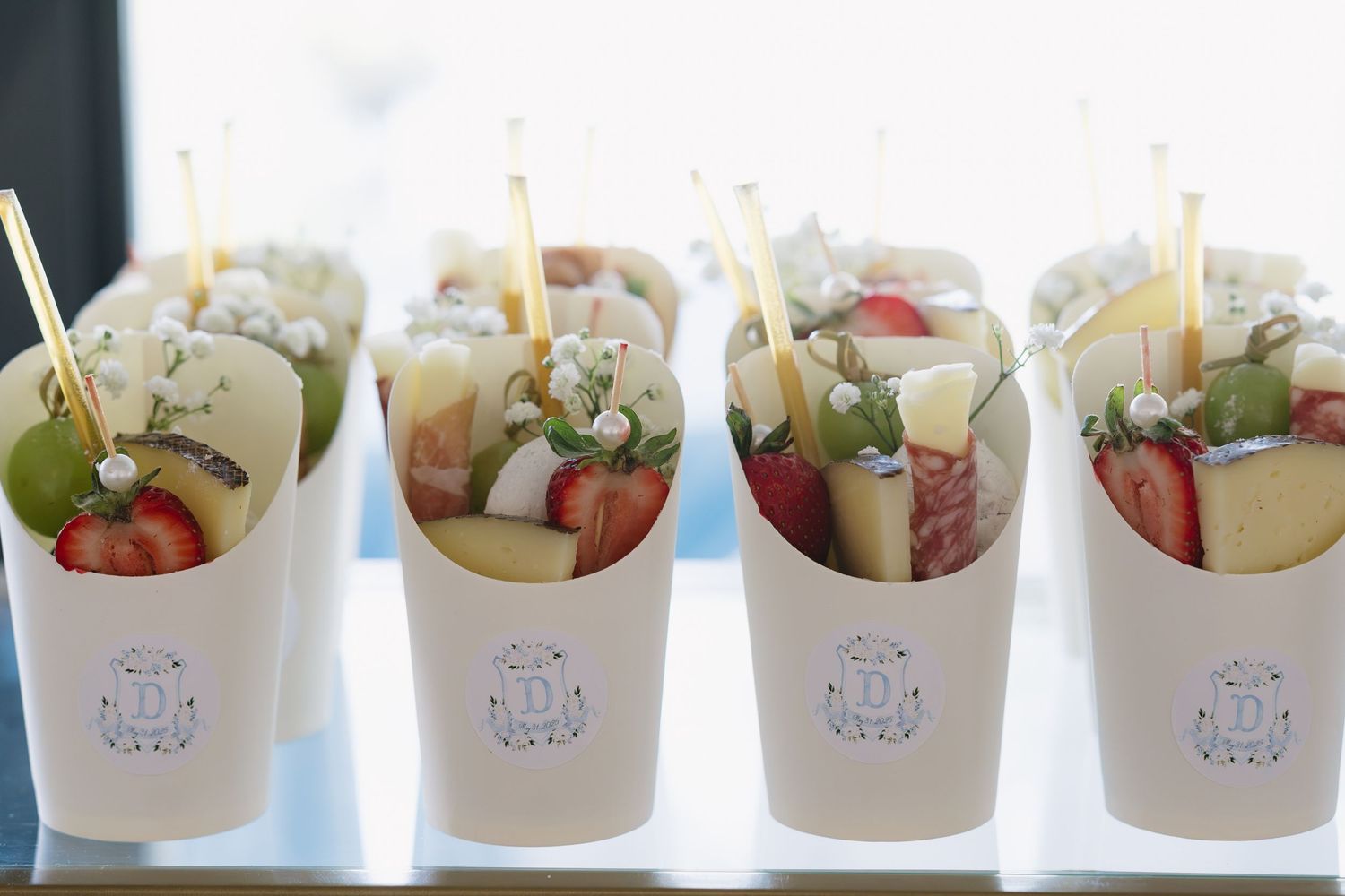 White paper cups filled with fresh fruit skewers including strawberries, grapes, and cheese, displayed in a row at a catered event.