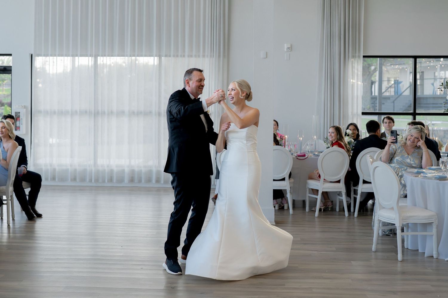 Bride and groom share their first dance in an elegant white wedding reception venue with sheer curtains and seated guests.