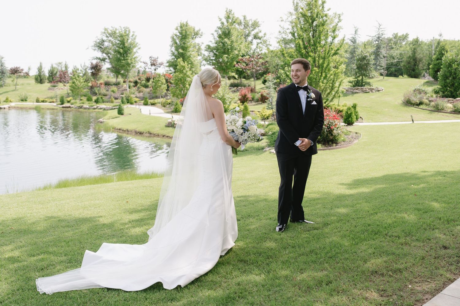 Bride in white gown with veil and groom in black tuxedo share a first look moment on lush green lawn by a pond.