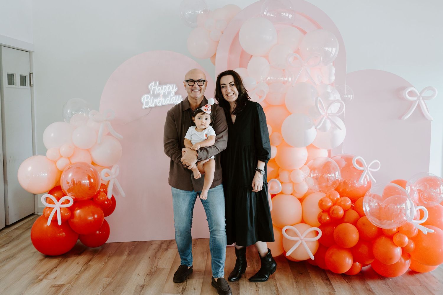 A couple poses with their baby against a backdrop of pink and orange balloons.