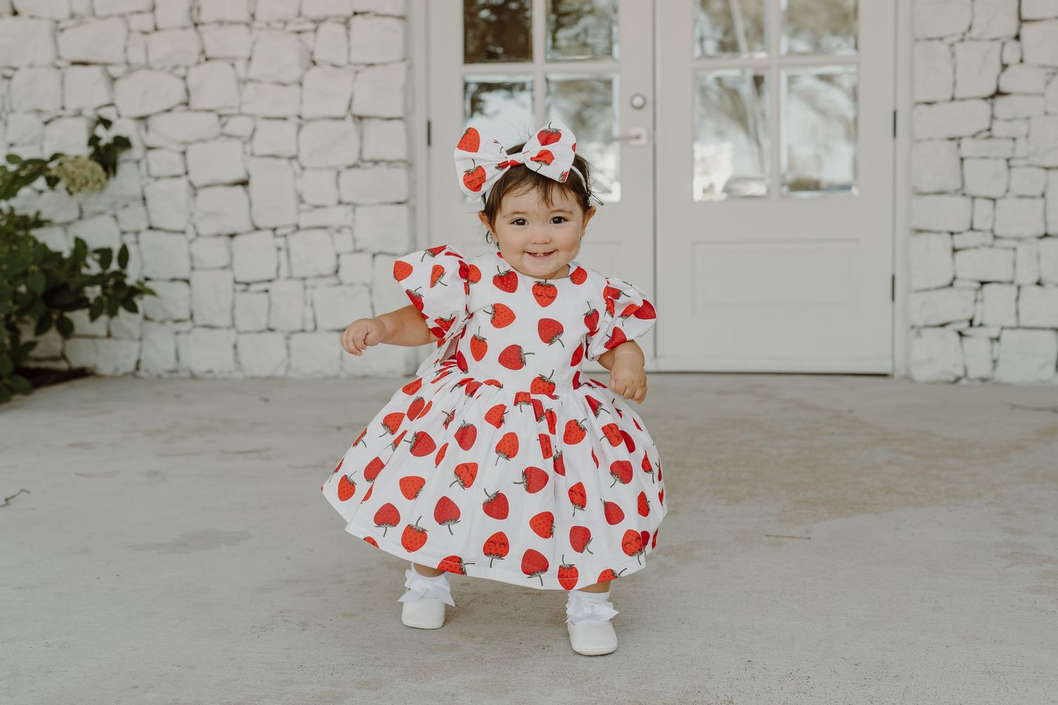 Child in heart-print dress twirling and playing against white brick wall.