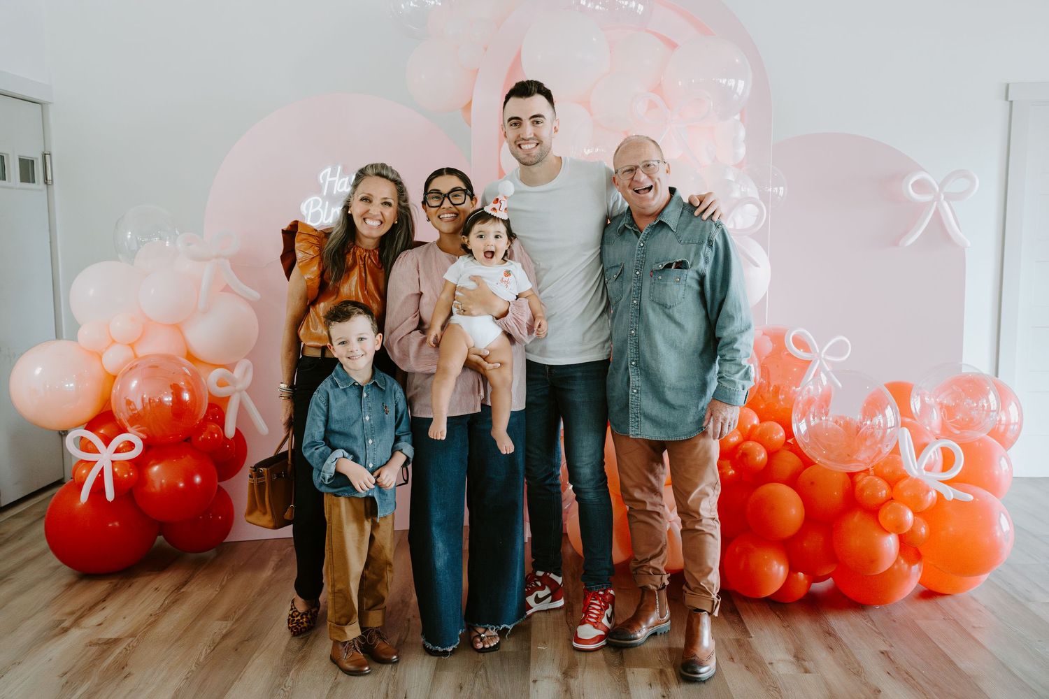 A group poses in front of a pink and orange balloon arch backdrop at a birthday celebration.