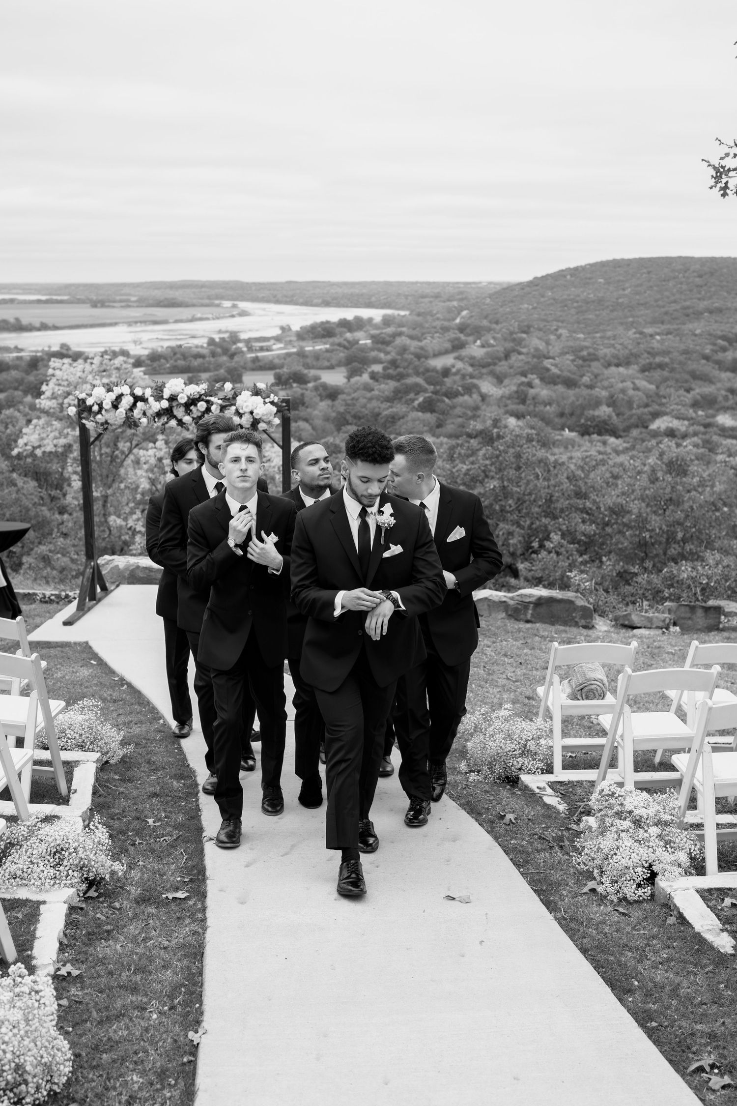 Wedding ceremony setup with white chairs and groomsmen walking along hillside path.