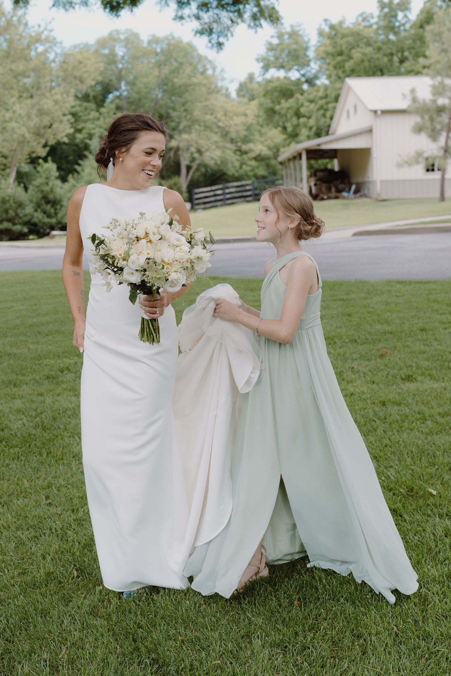 Two people in white and mint green dresses sit together on grass holding bouquets during a wedding.