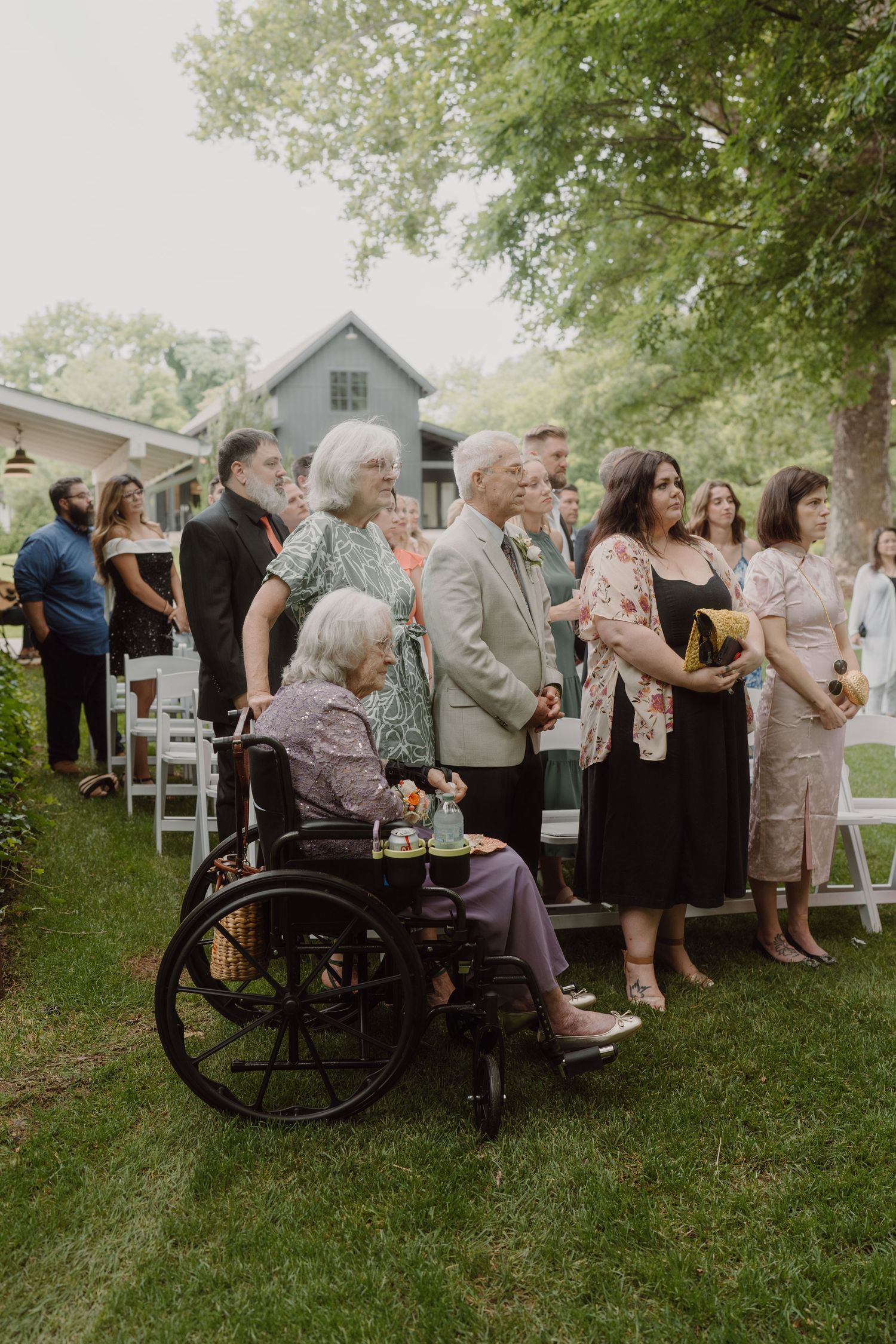 Wedding guests, including some in wheelchairs, attend an outdoor ceremony on a grassy lawn.