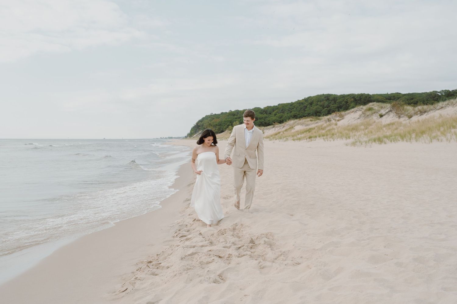 A couple strolls hand in hand on a scenic beach with green dunes in the distance.
