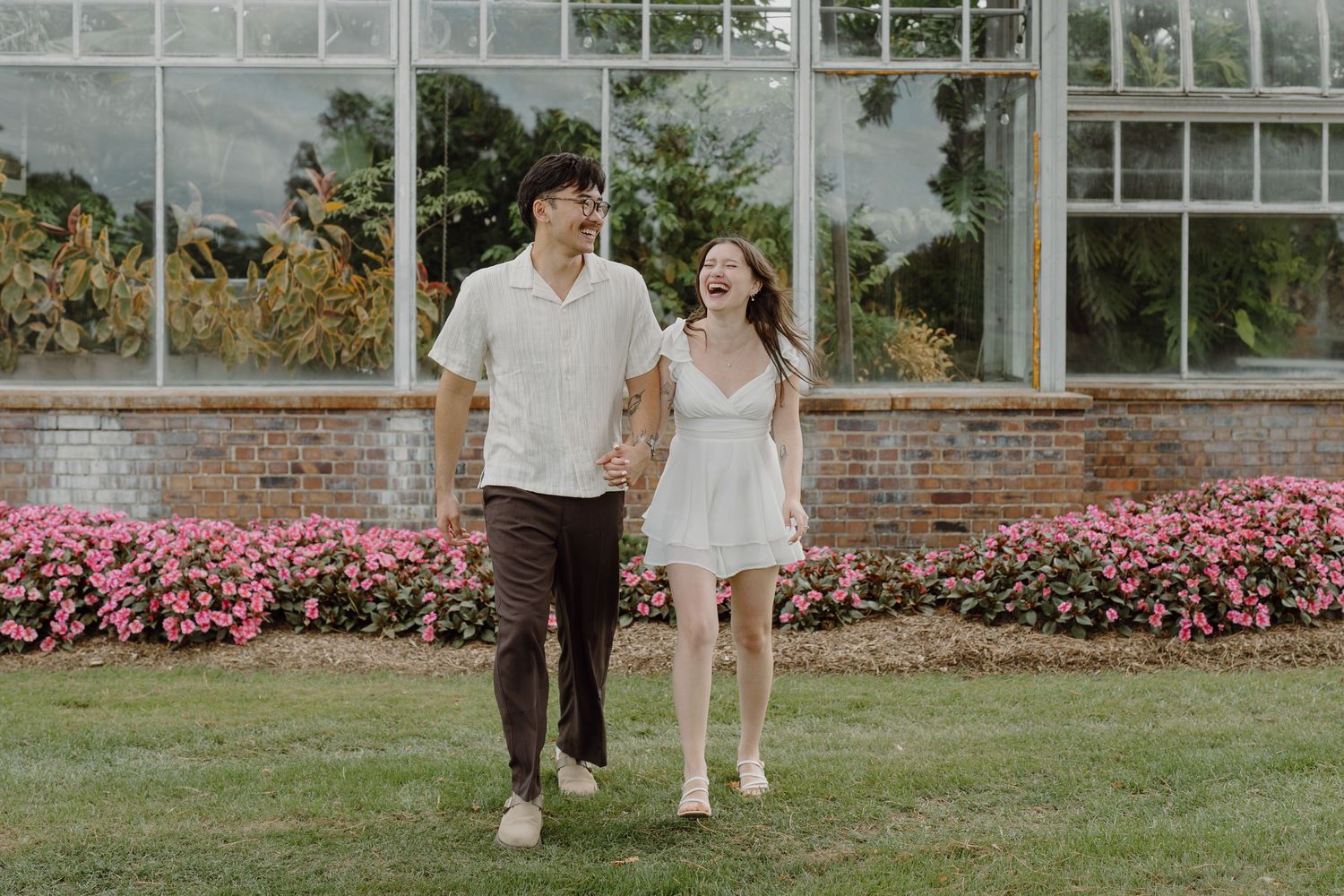 Two people pose together on grass near a brick wall and greenhouse with pink flowers.