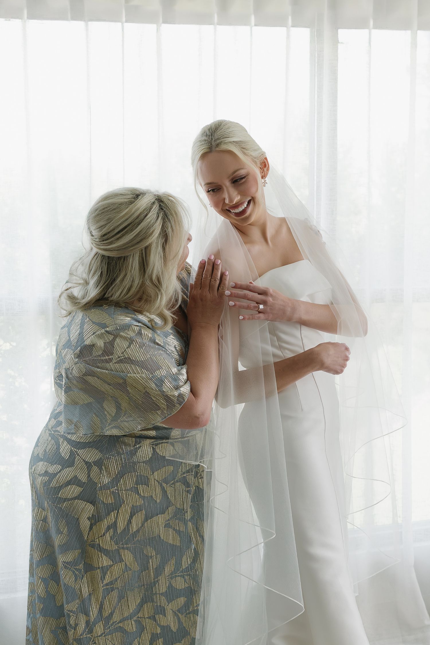 A smiling bride in a white gown and veil shares a joyful moment with an older woman in a floral dress.