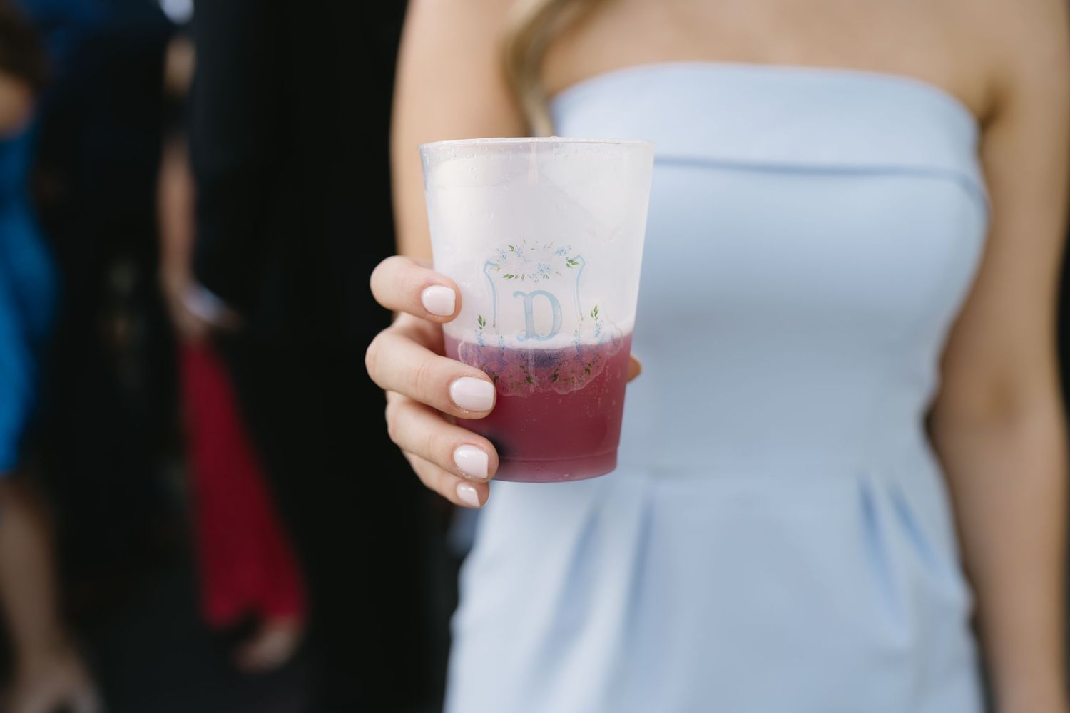 Woman in light blue strapless dress holding a frosted monogrammed cup with red drink at a wedding.