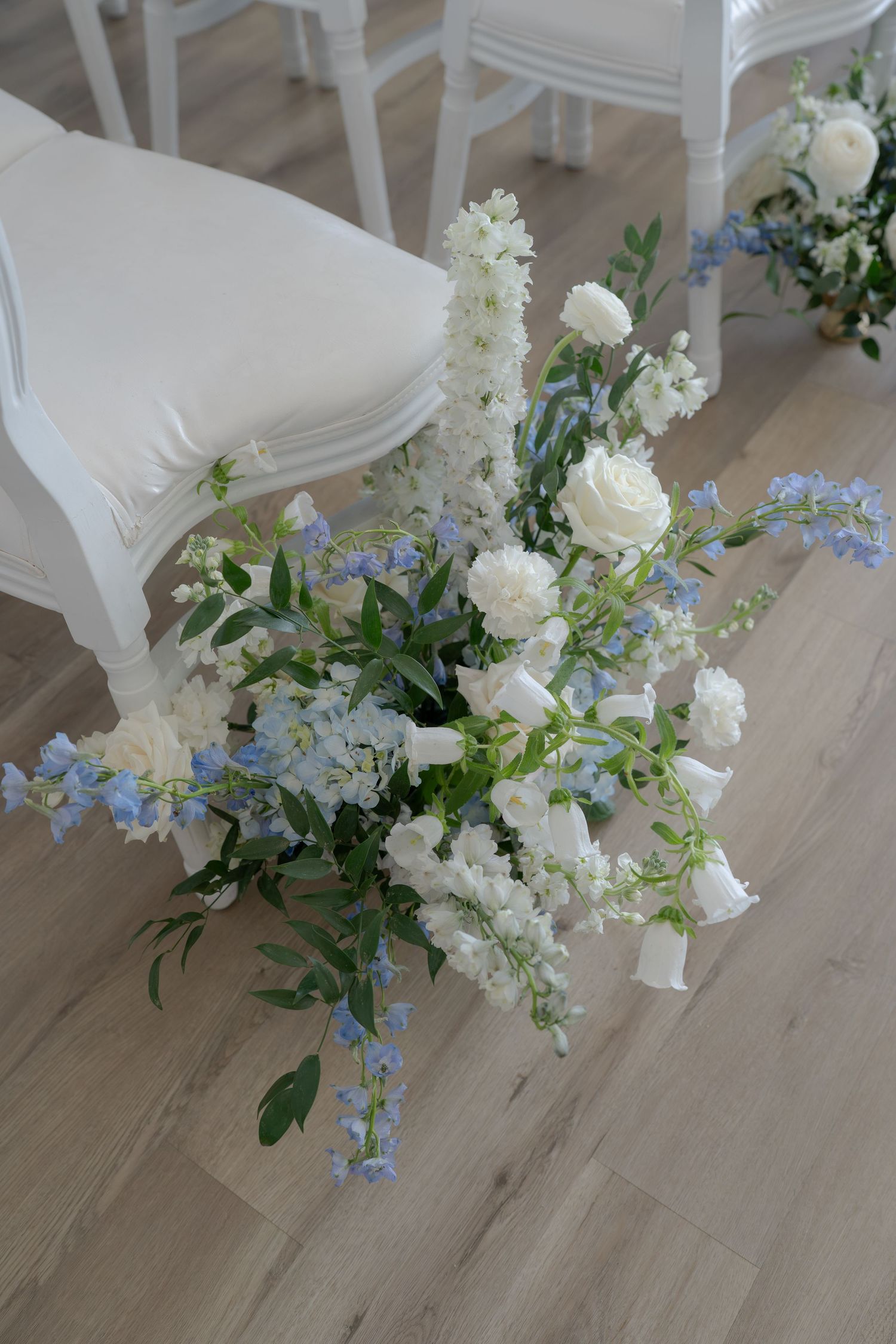 White and blue floral arrangement with roses and delphinium beside elegant white chairs at a wedding ceremony.