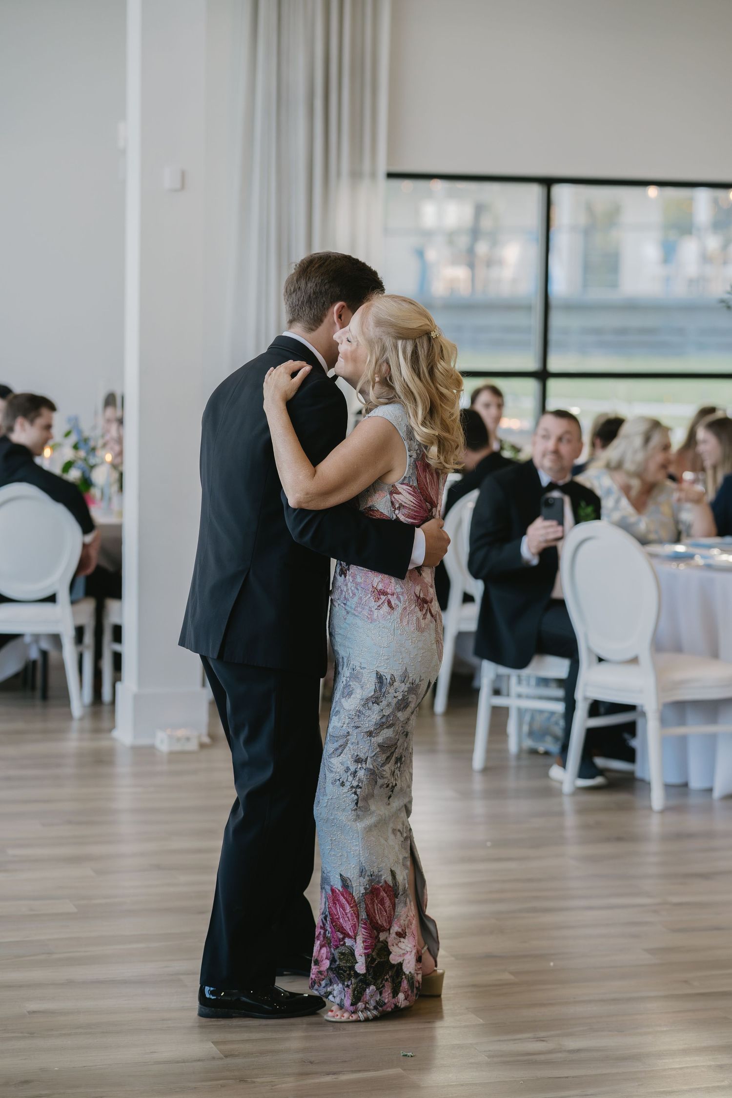 Couple shares romantic first dance at elegant wedding reception, woman in floral gown, guests seated at white chairs.