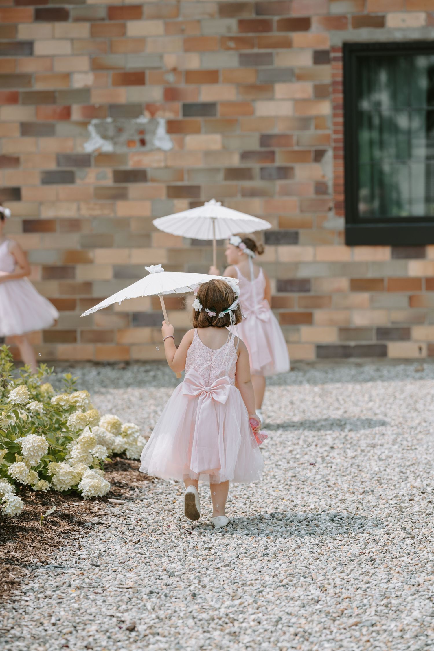 Young flower girls with parasols walk along a gravel path near a brick building.