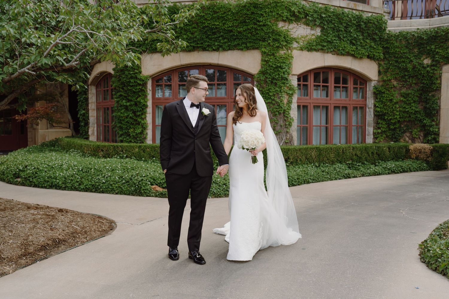 A wedding couple walks along a curved garden path surrounded by lush greenery and brick buildings.