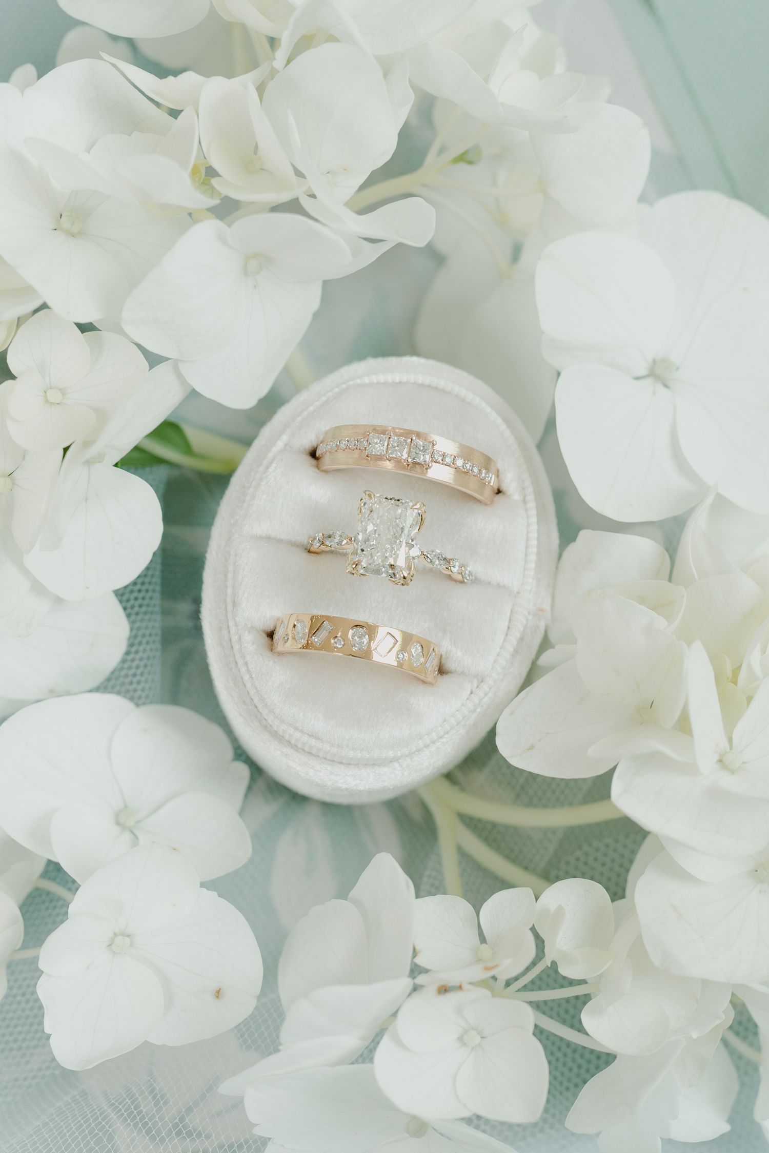 Three gold diamond wedding rings displayed in a white velvet ring box surrounded by white hydrangea flowers.