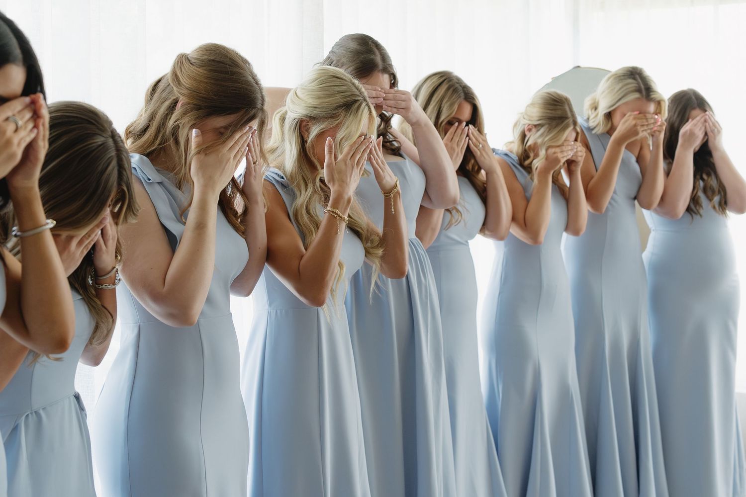 Bridesmaids in matching light blue dresses stand in a row, covering their faces with their hands.