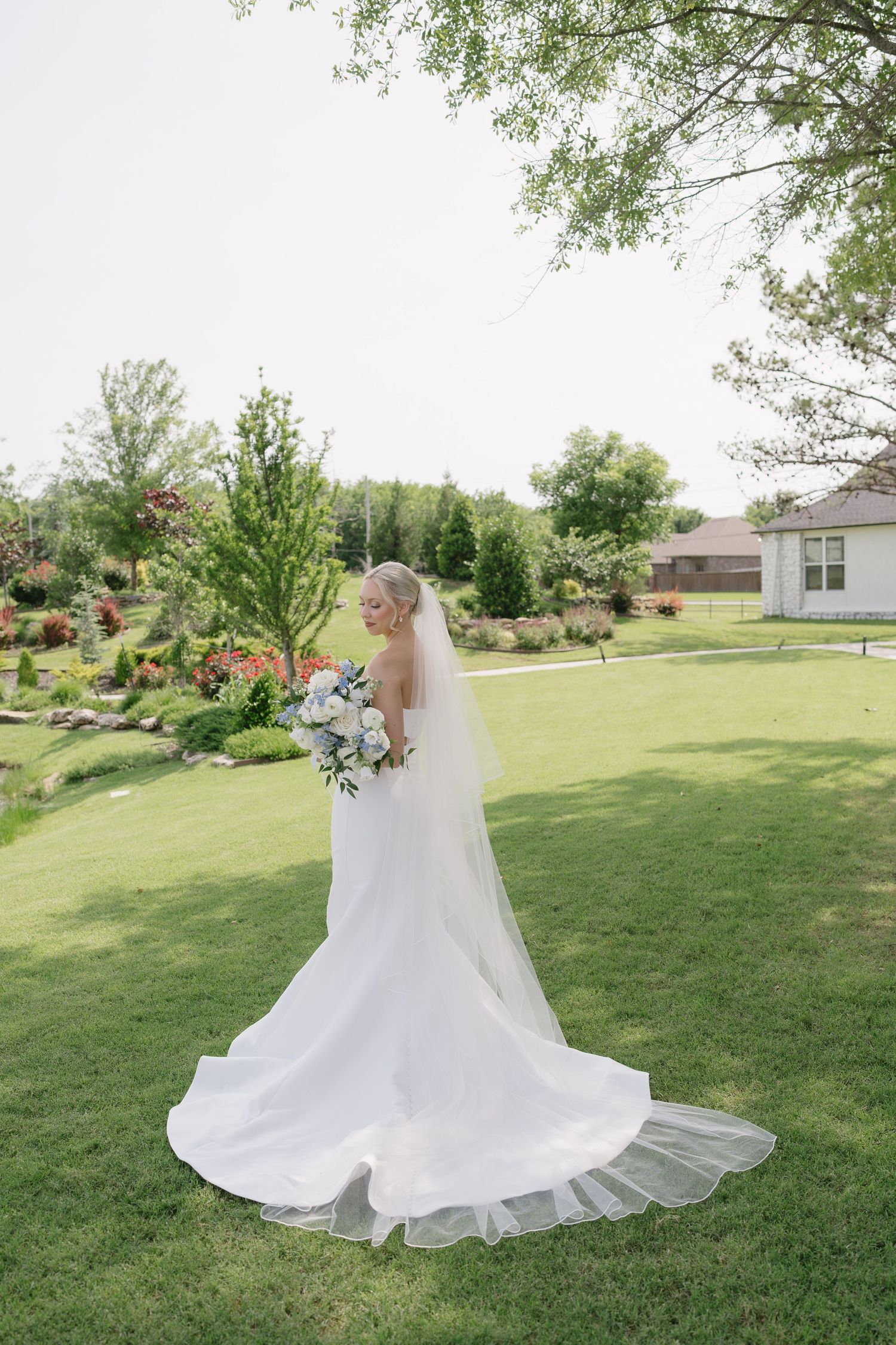 Bride in white gown with long veil holds bouquet on green lawn surrounded by lush garden trees.