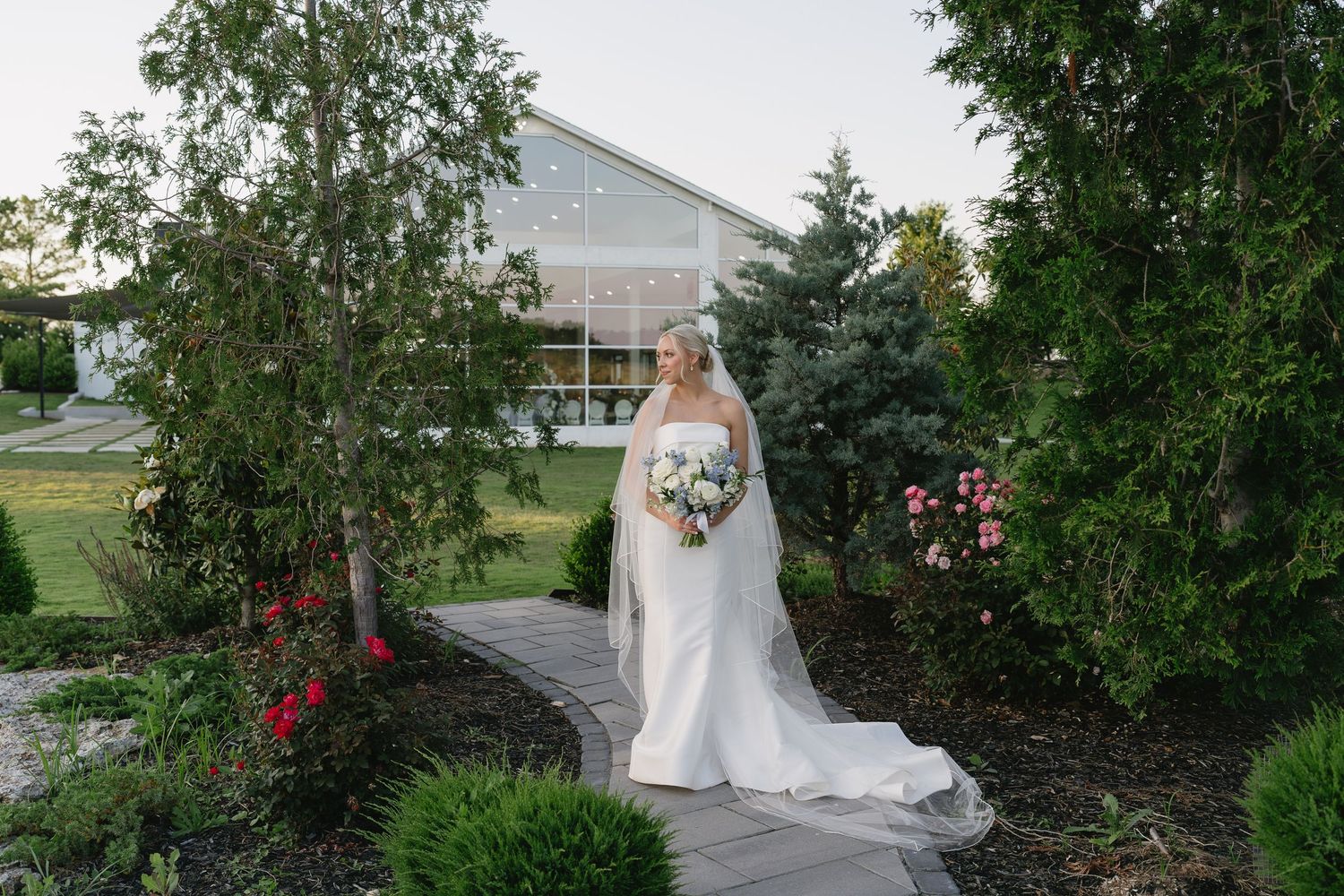 Bride in white gown and veil holding bouquet on stone garden path with lush greenery and venue in background.