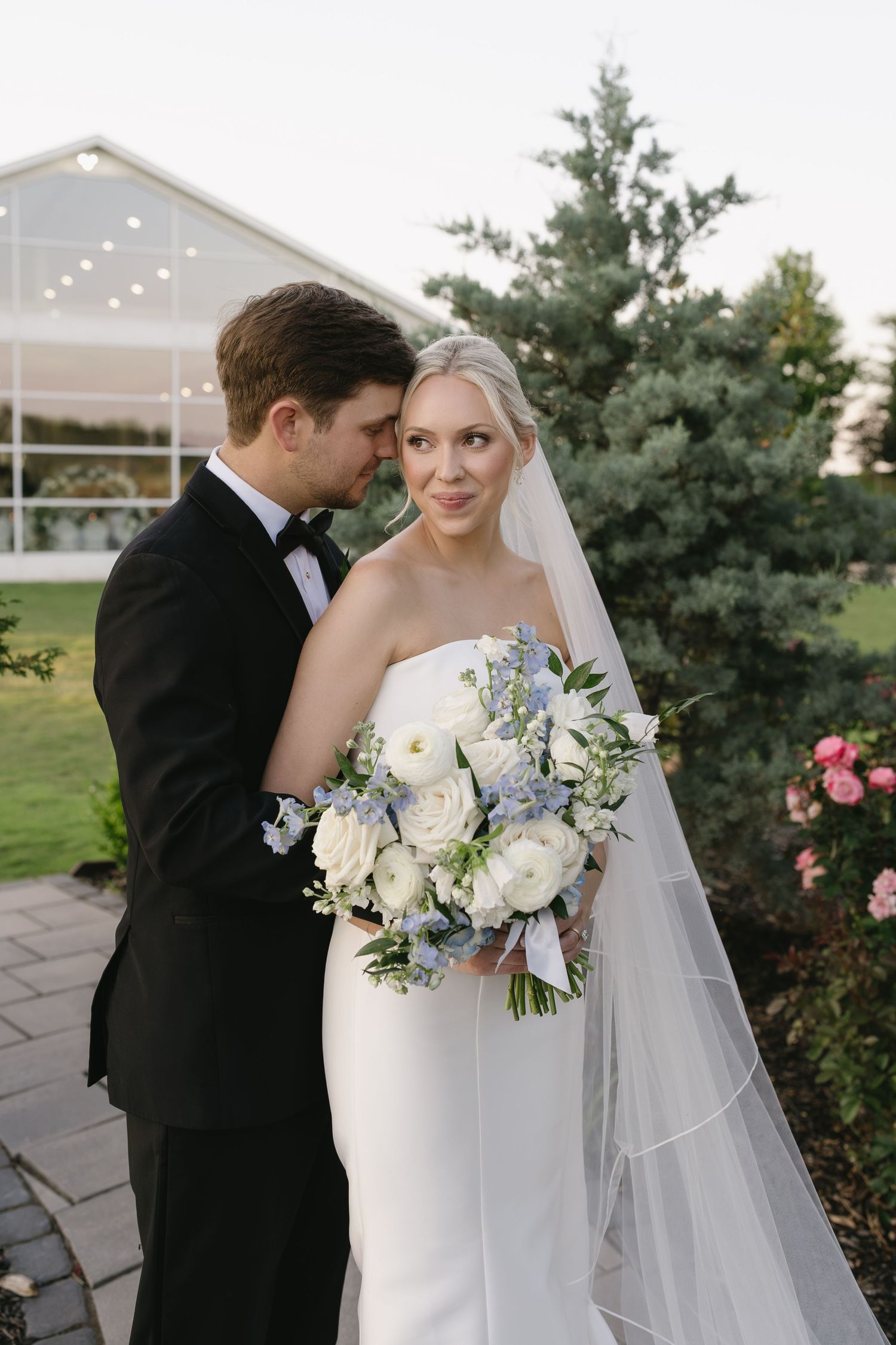 Bride in white strapless gown holding blue and white bouquet stands with groom in black tuxedo in garden setting.