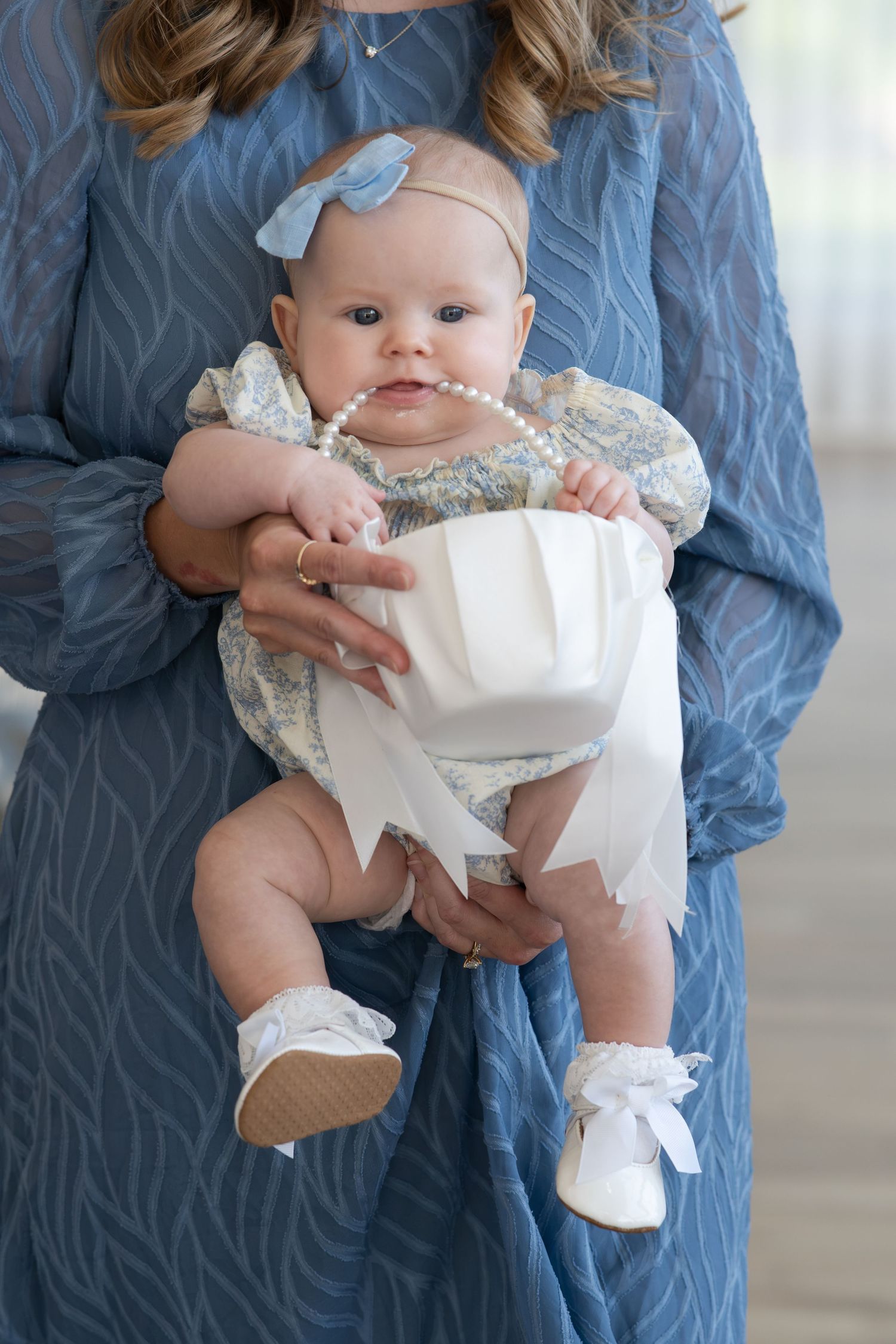 A baby girl in white christening outfit with pearl necklace, held by a woman in blue dress.