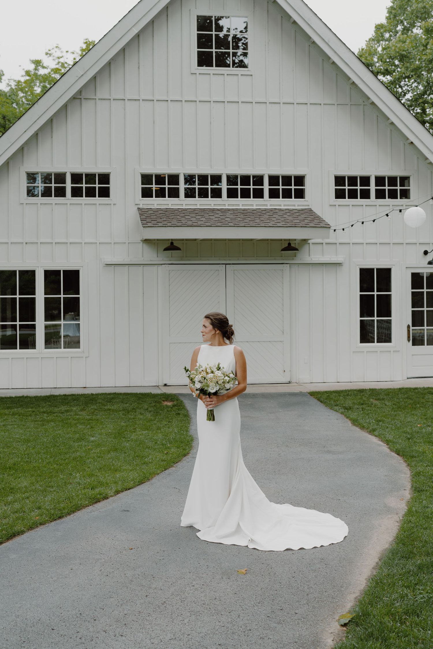A bride in a sleek white gown stands on a curved path near a white barn-style venue.