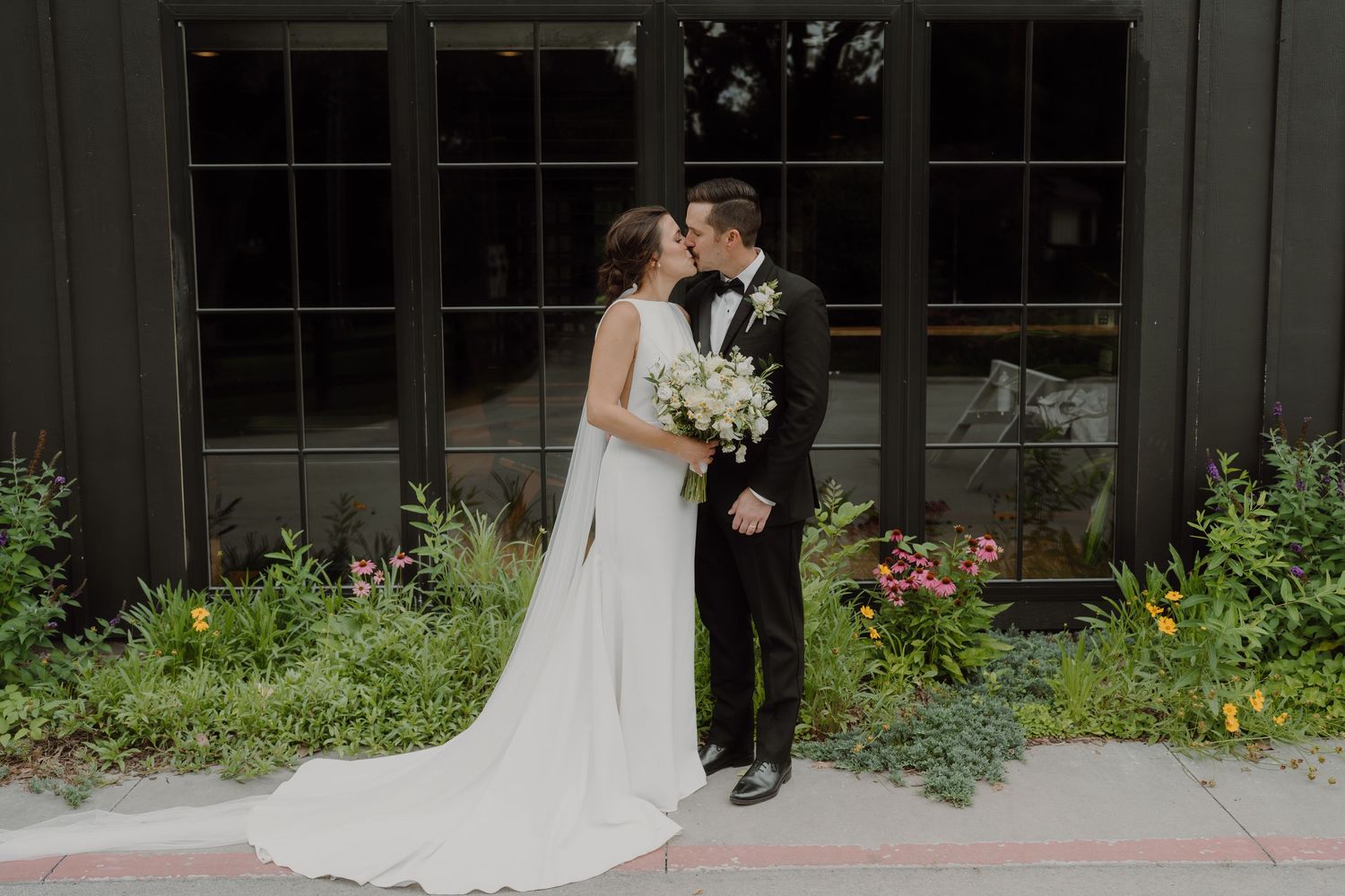 Elegant wedding couple poses against Spain Ranch's modern black barn venue with large windows.