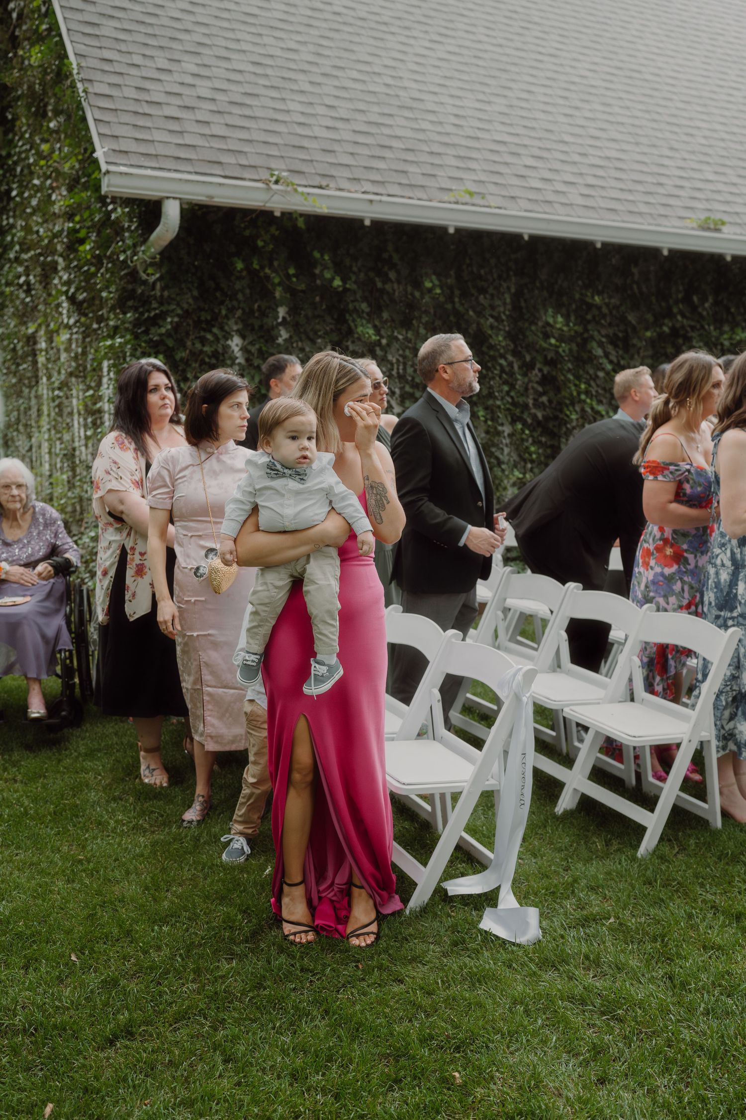 Wedding guests in colorful attire stand beside white folding chairs during an outdoor ceremony.