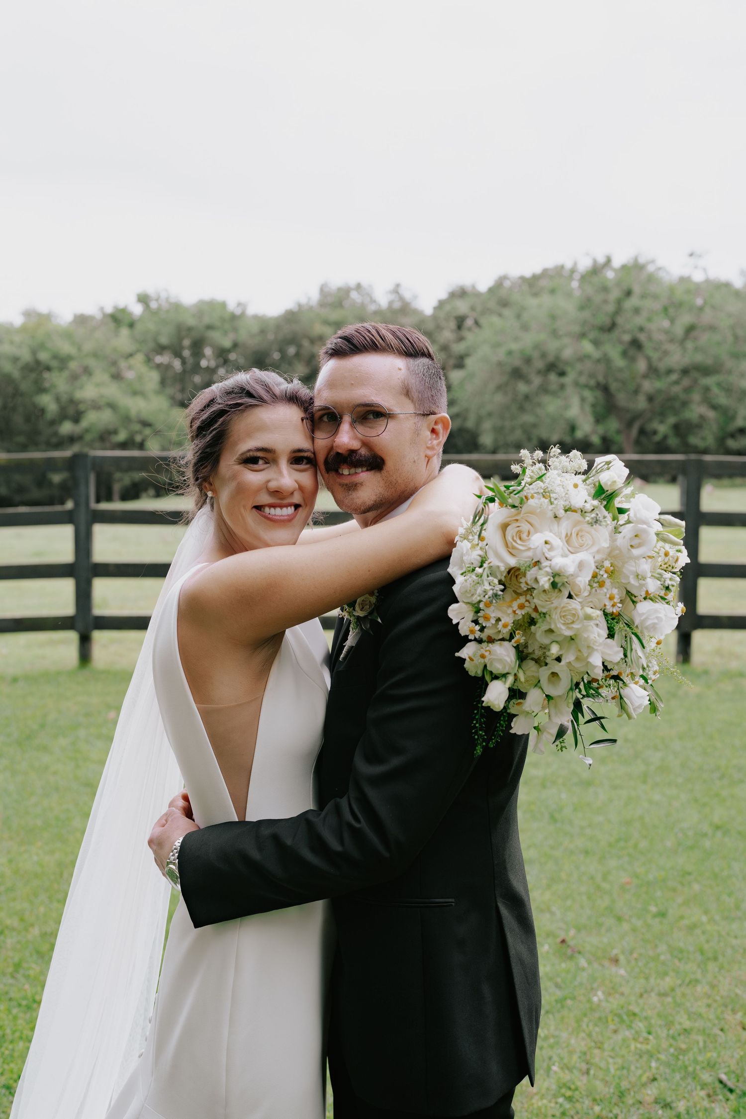 A romantic wedding photo series showing a couple embracing in a grassy field with white fencing and trees in the background.