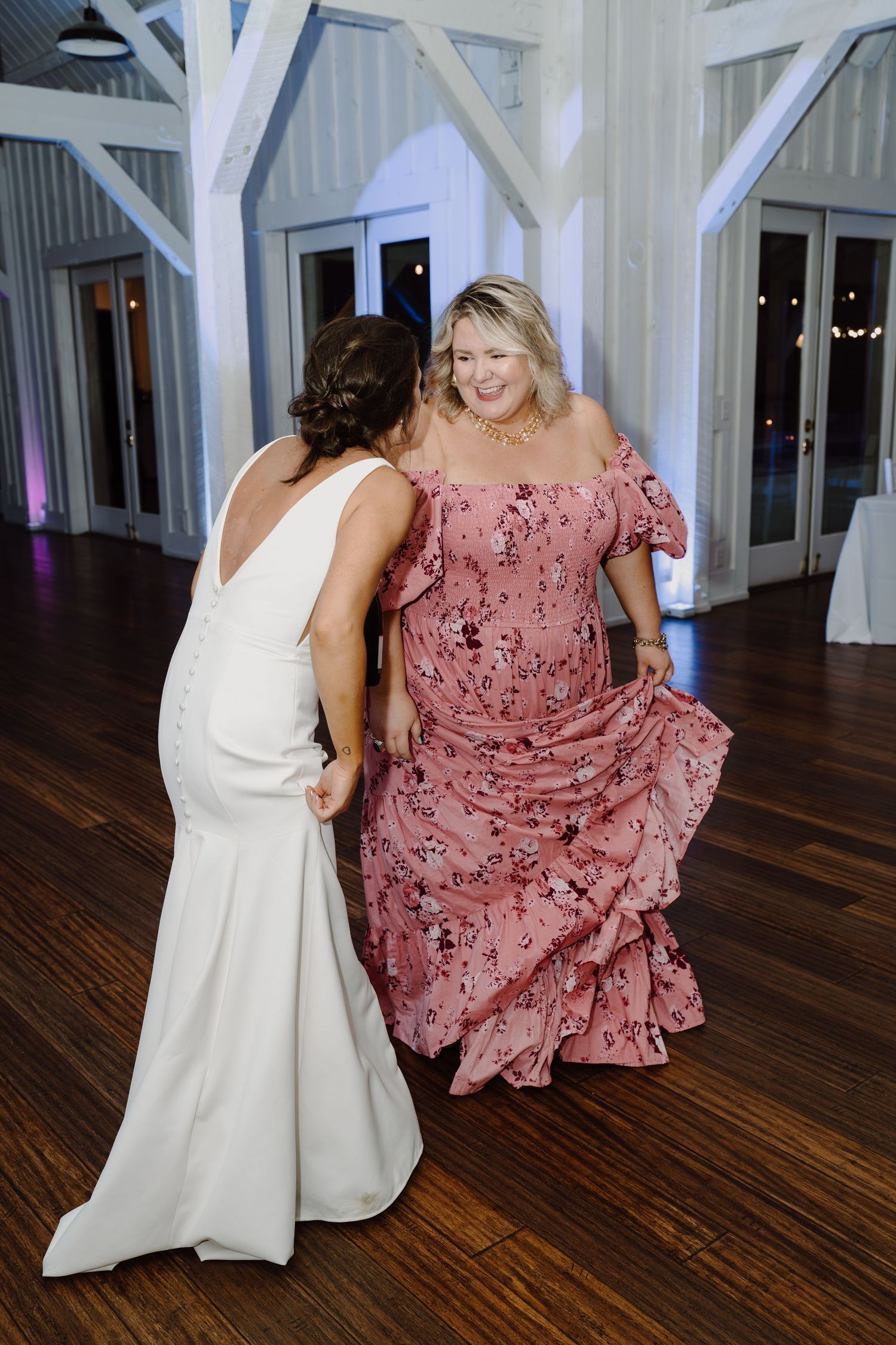 Two women dancing together at a wedding reception, one in white and one in pink floral dress.