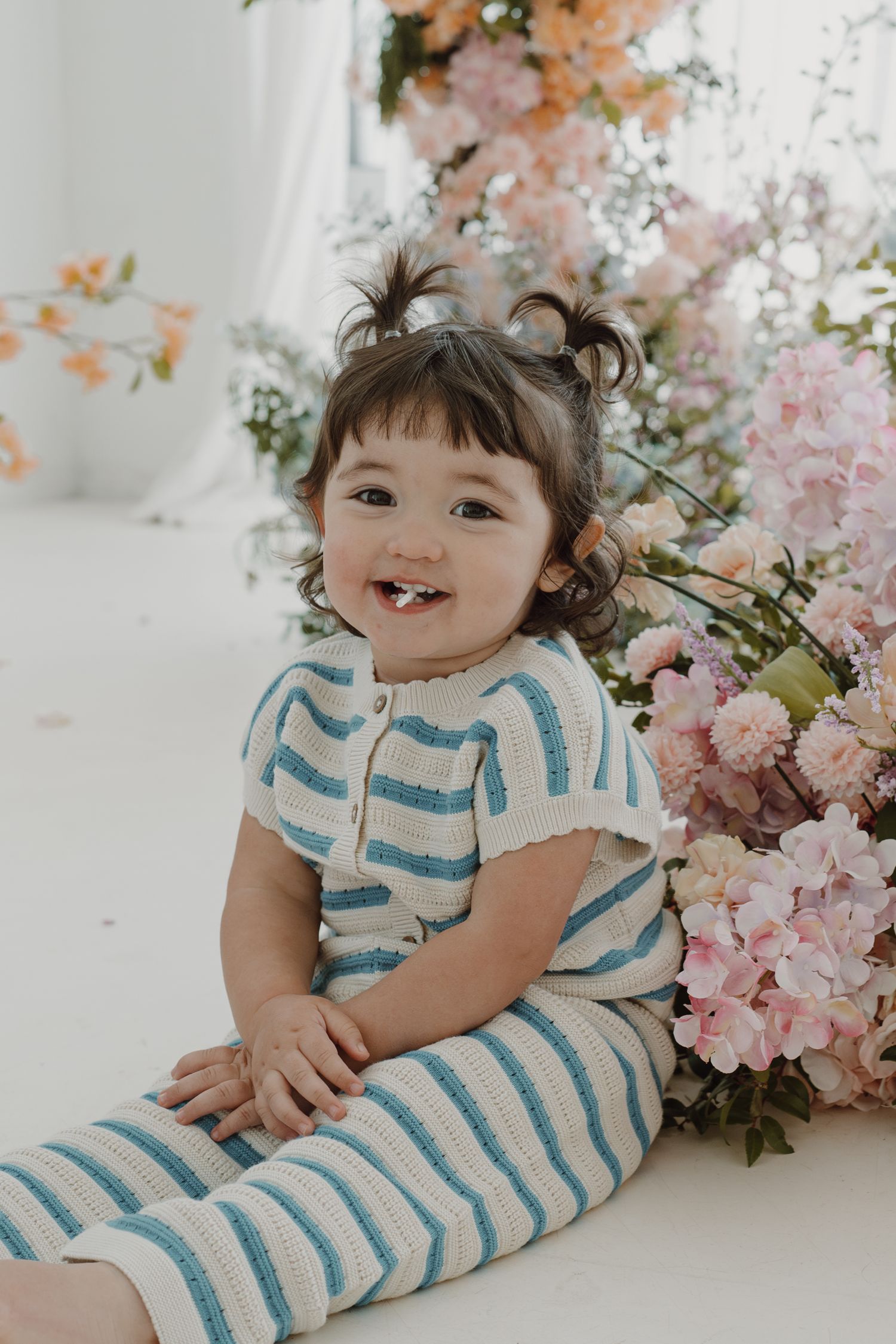 Happy toddler in striped outfit sitting by pink floral arrangements in bright studio setting.