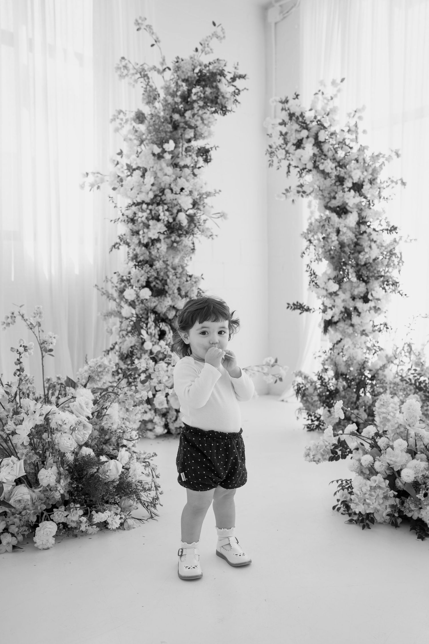 Black and white photo of toddler in studio with elegant floral backdrop.
