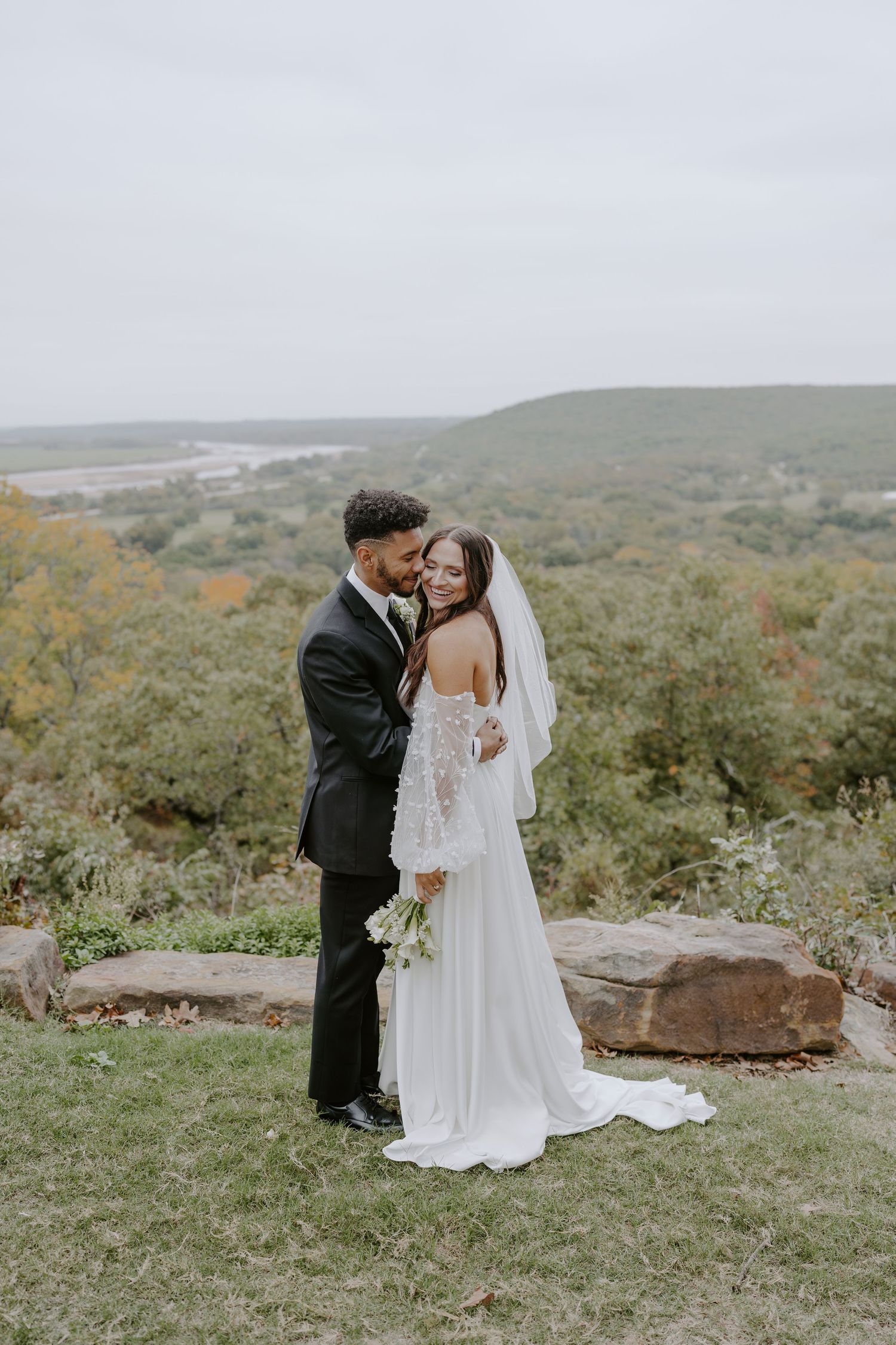 A wedding couple shares an intimate moment on a scenic overlook with autumn foliage in the background.