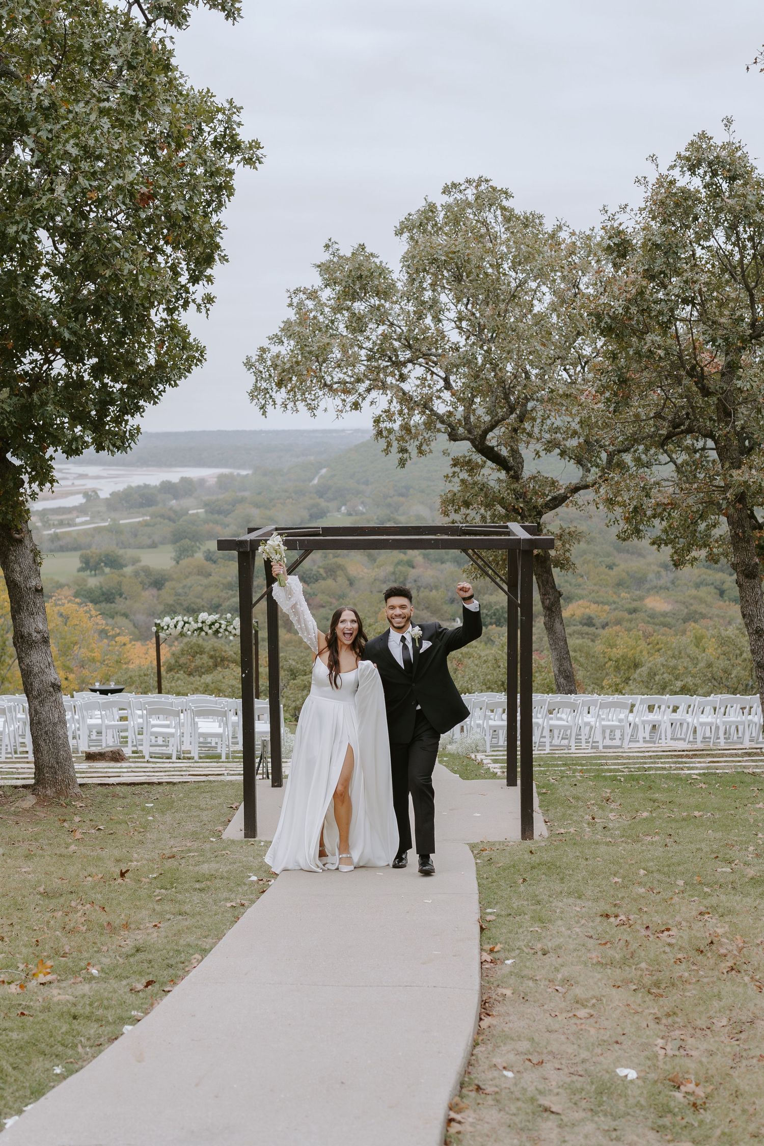 A wedding couple poses under a wooden arch on a hilltop overlooking a scenic valley with rows of white chairs set up.